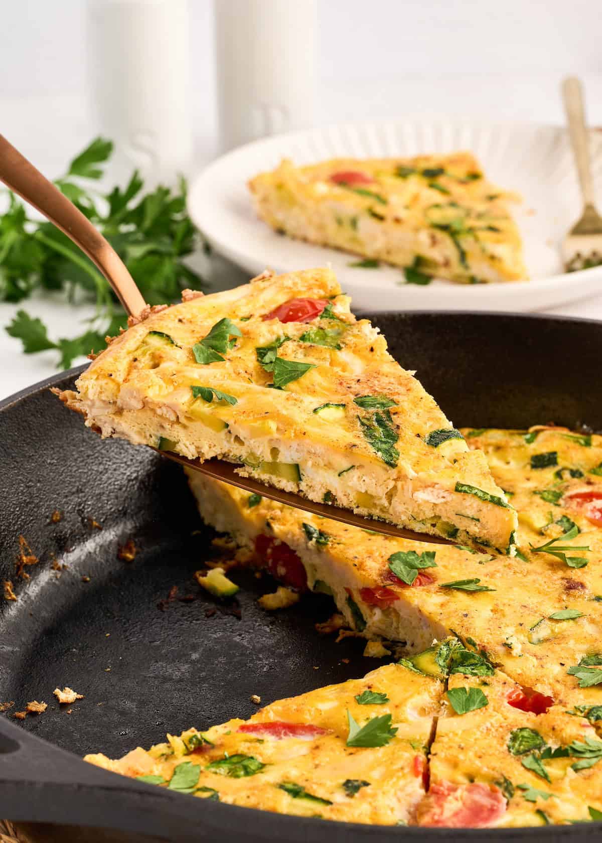 Wedge of frittata with shredded chicken, tomatoes, zucchini, and herbs in a cast iron skillet.