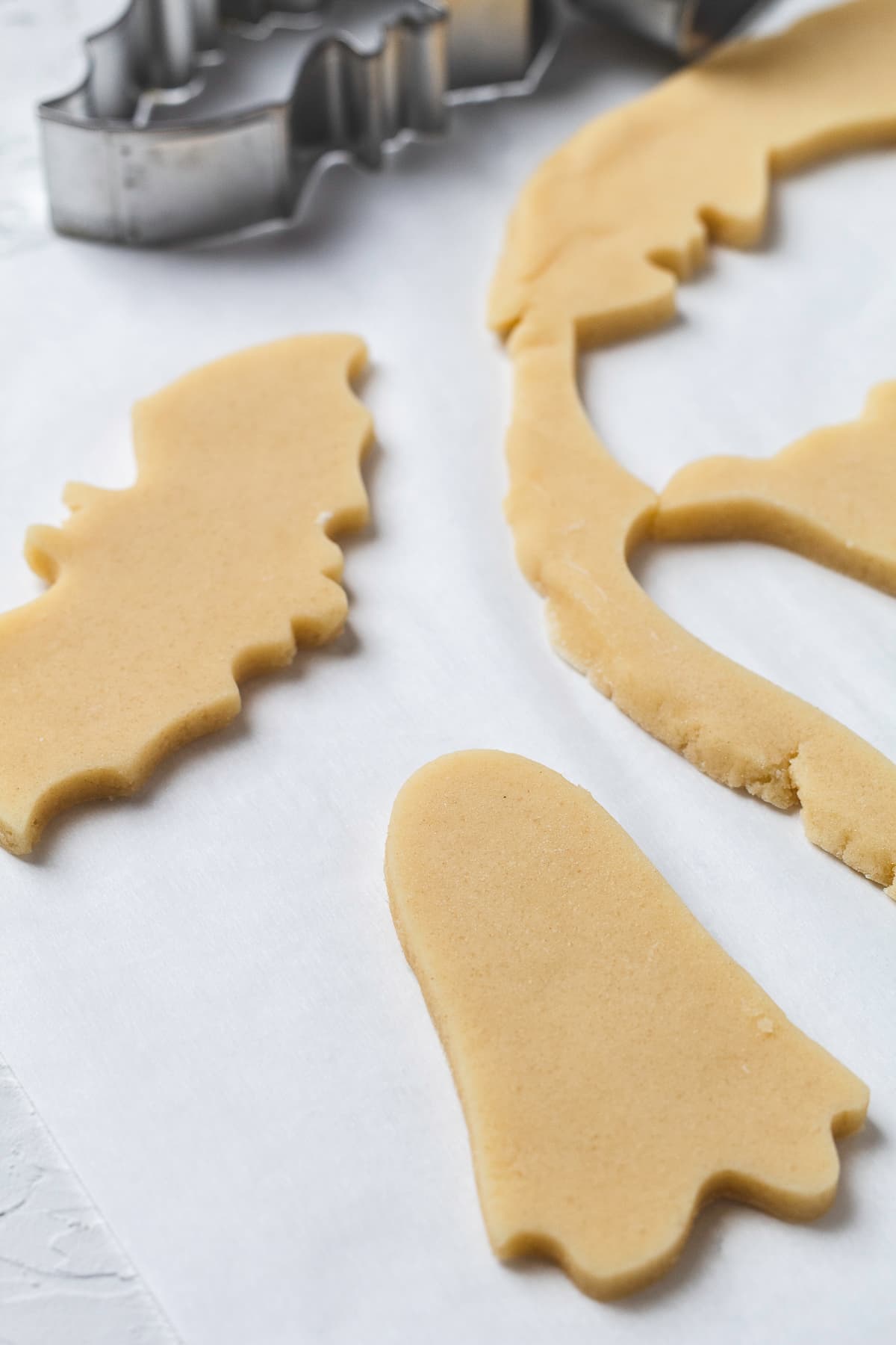 Unbaked ghost and bat-shaped cookies cut from dough, resting on parchment paper.