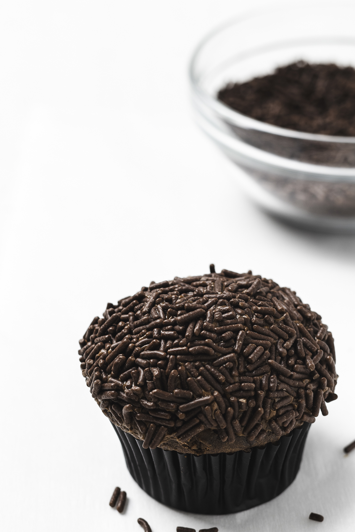 Chocolate frosted cupcake covered with chocolate sprinkles with more in the background.