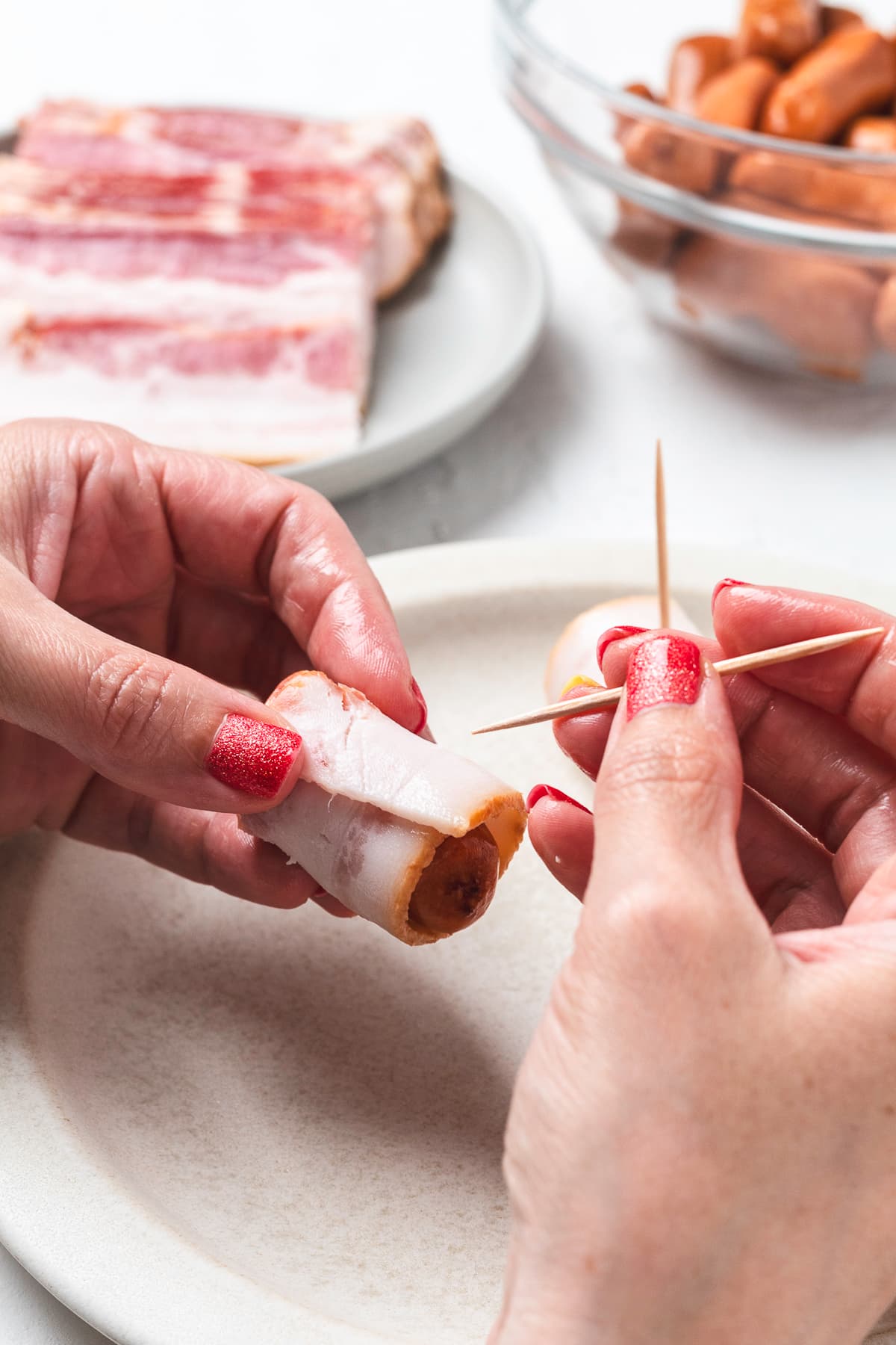 Hands inserting a toothpick into bacon-wrapped sausage.