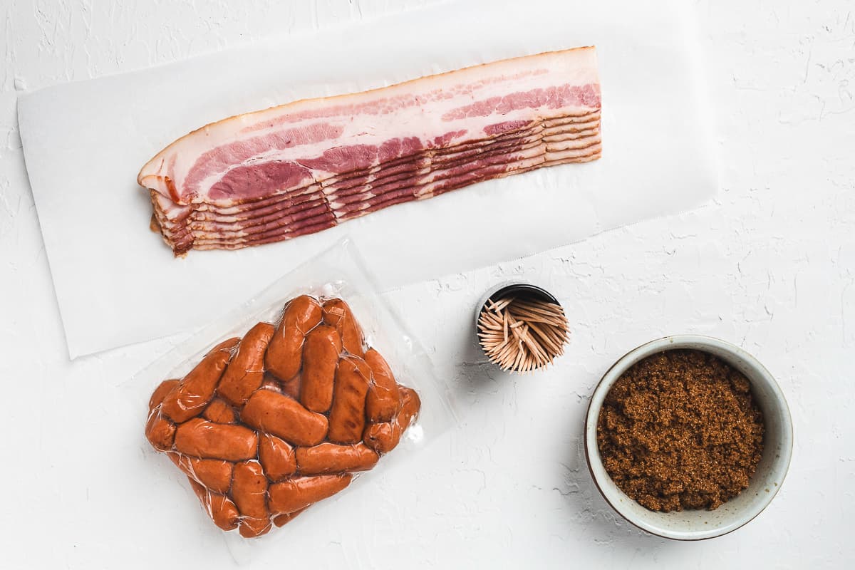 Ingredients for bacon-wrapped little smokies including bacon, sausages, toothpicks, and brown sugar.