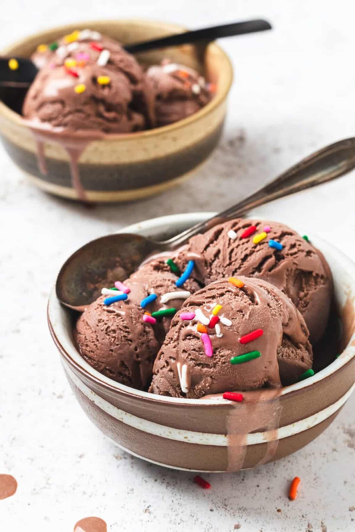 Two bowls of chocolate ice cream with spoons on a light colored background.
