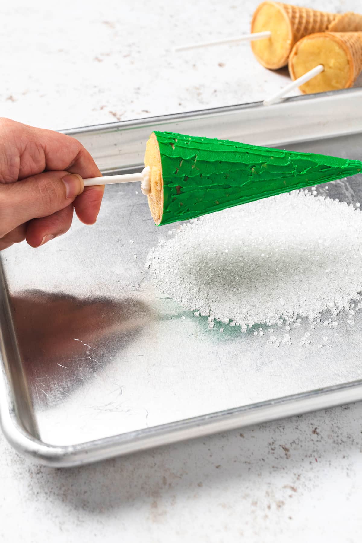 Hand holding Christmas tree cake pop ready to sprinkle decorative sugar on it.