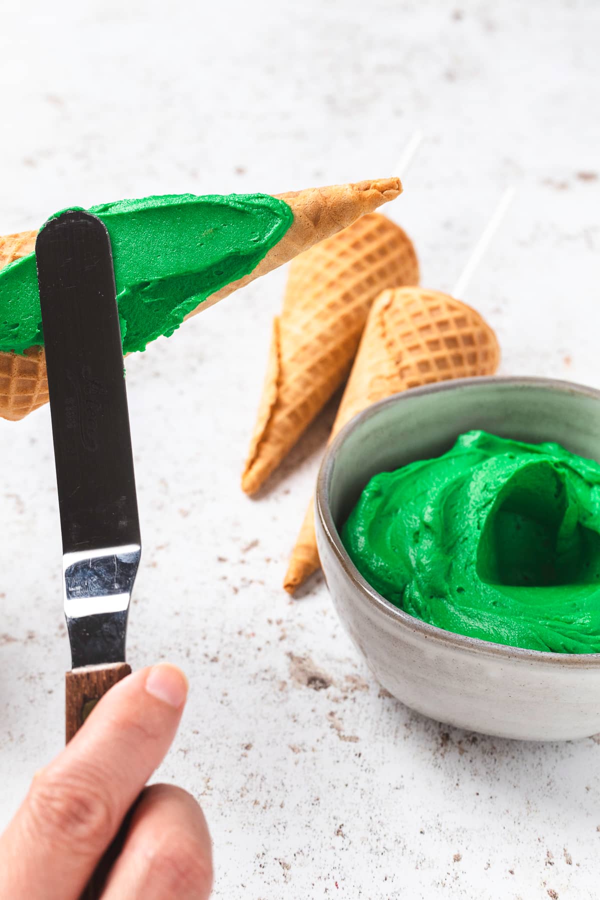 Hand spreading green frosting on a sugar cone.