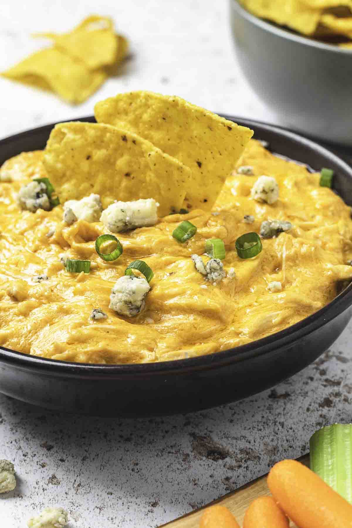 Buffalo chicken dip in a dark brown bowl with tortilla chips, blue cheese, and green onions.