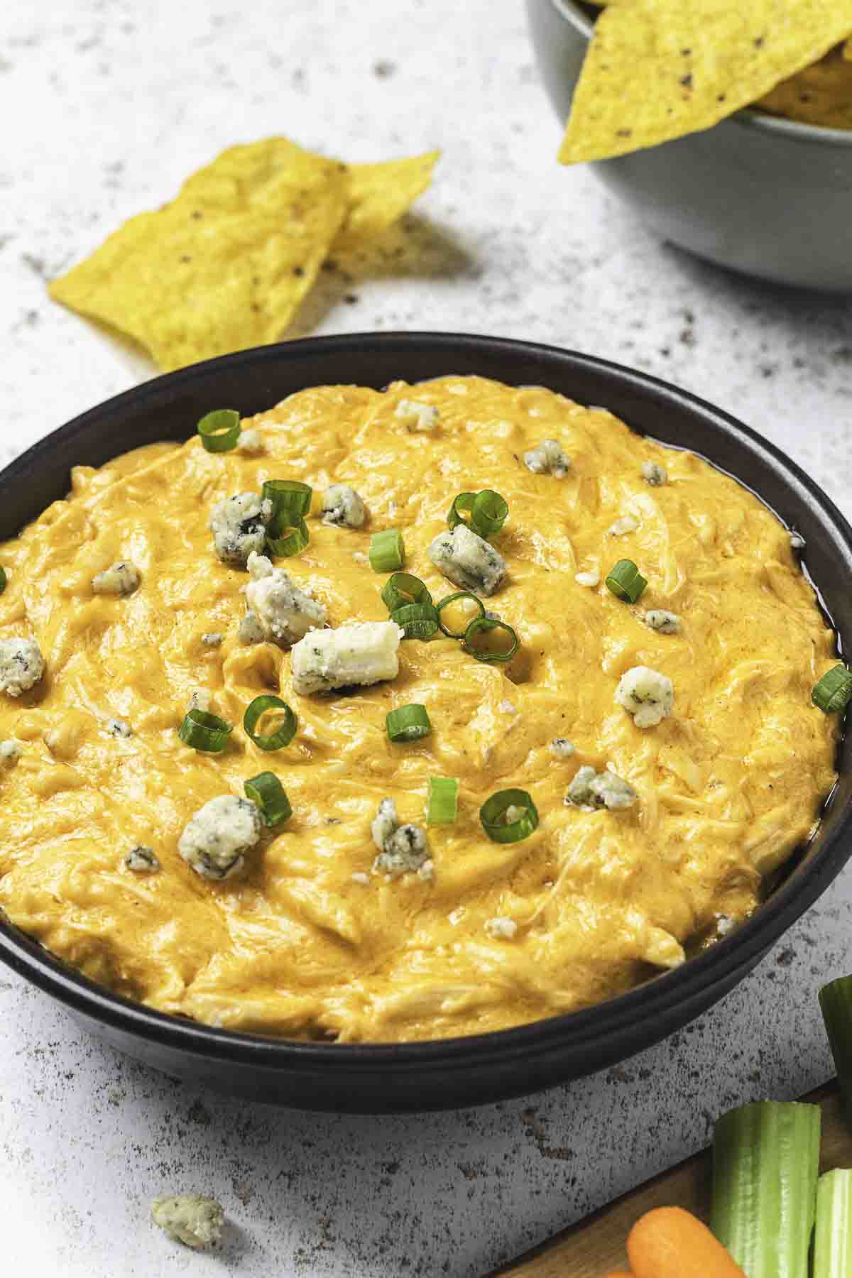 Buffalo chicken dip in brown bowl with blue cheese, green onions, and dippers on a light colored background.