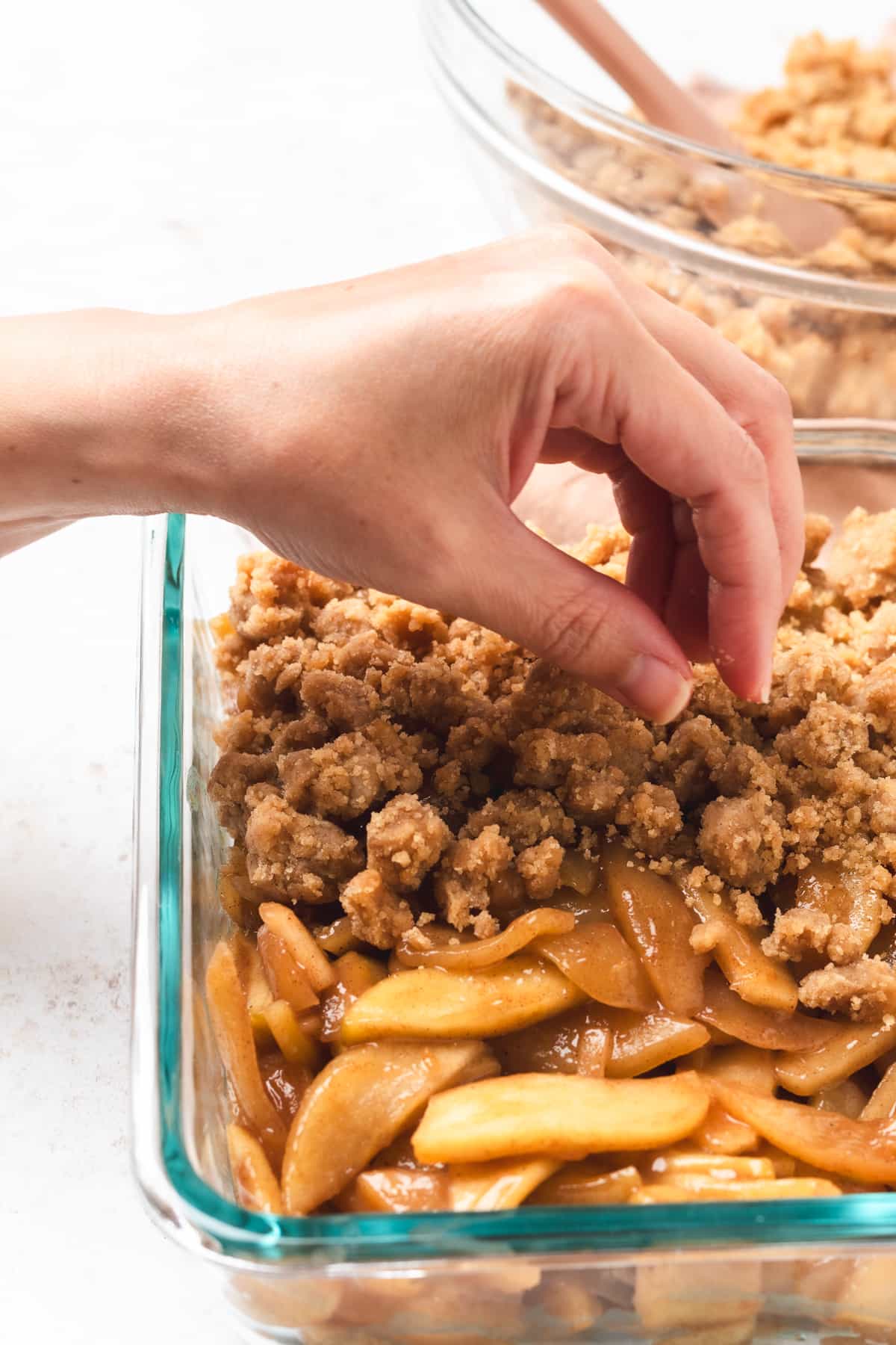 Hand sprinkling crumble topping over sliced apples in a glass baking dish.