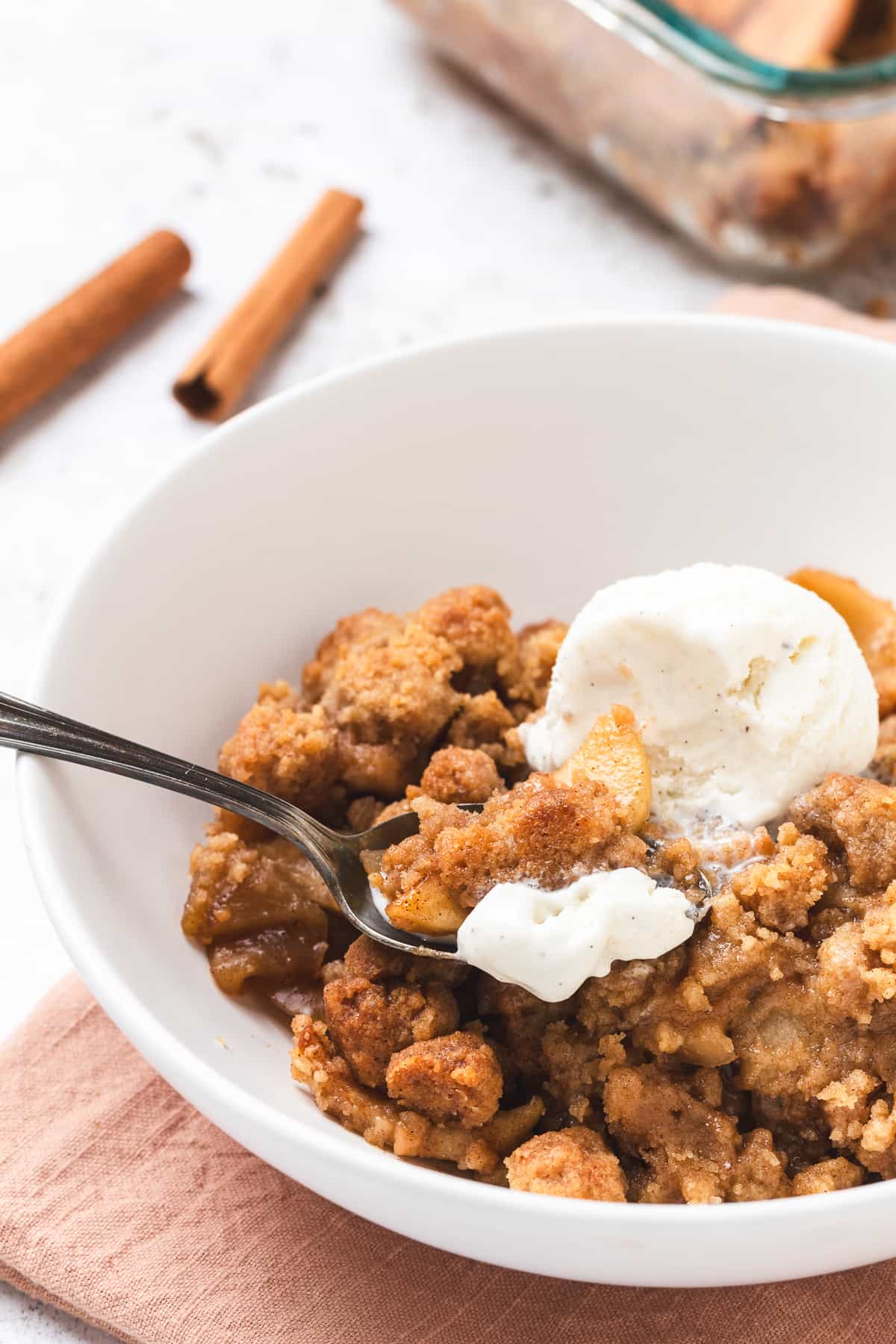 Bowl of apple crumble with ice cream and a spoon, cinnamon sticks in the background.