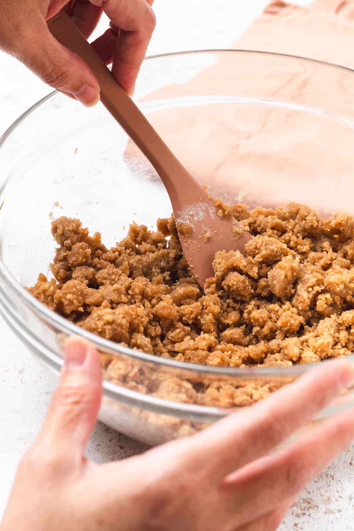 Mixing apple crumble topping in a clear glass bowl with a spatula.