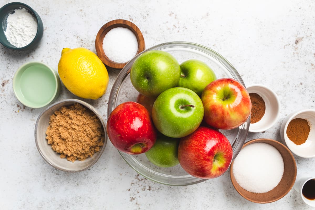 Fresh apples, lemon, sugar, spices, and other apple pie filling ingredients on a counter.
