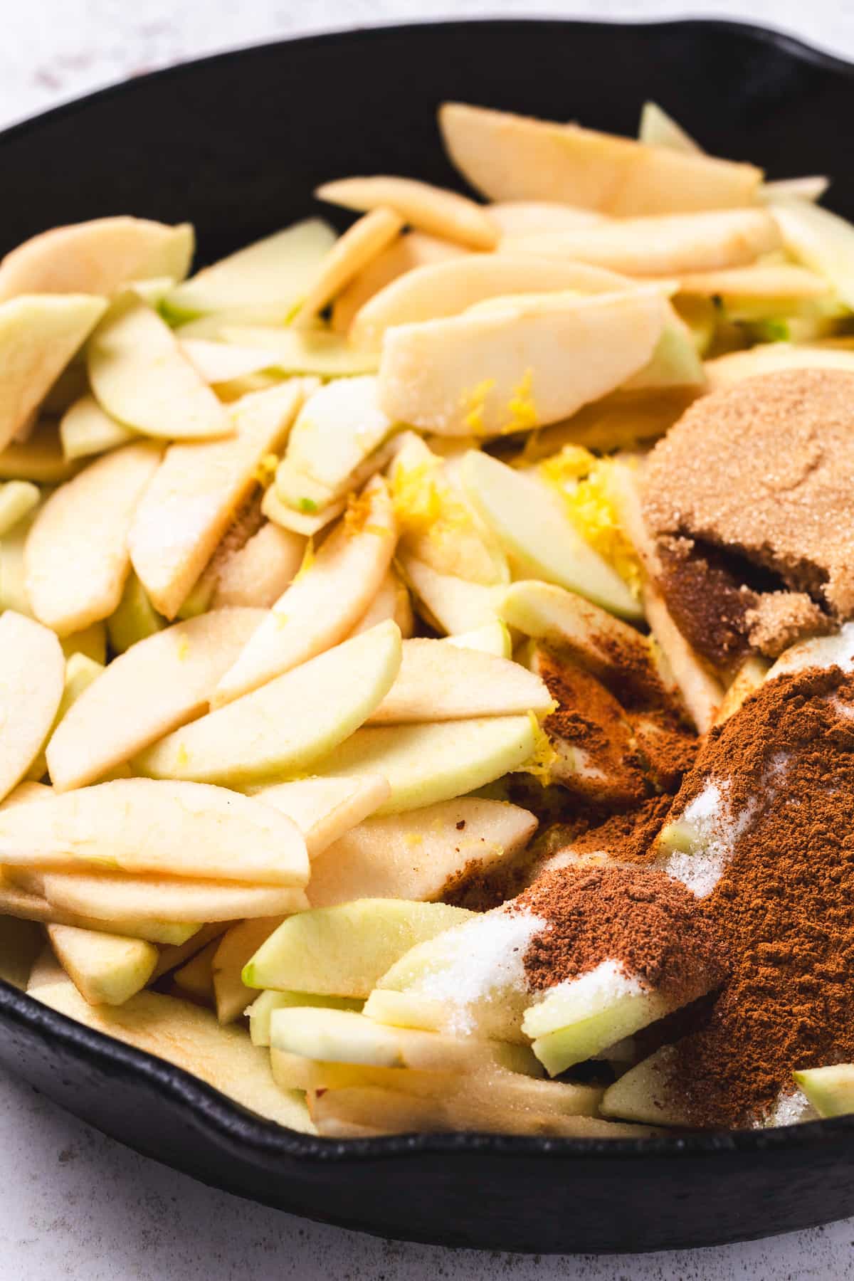 Sliced apples with sugar, brown sugar, cinnamon, and spices in a cast iron skillet.
