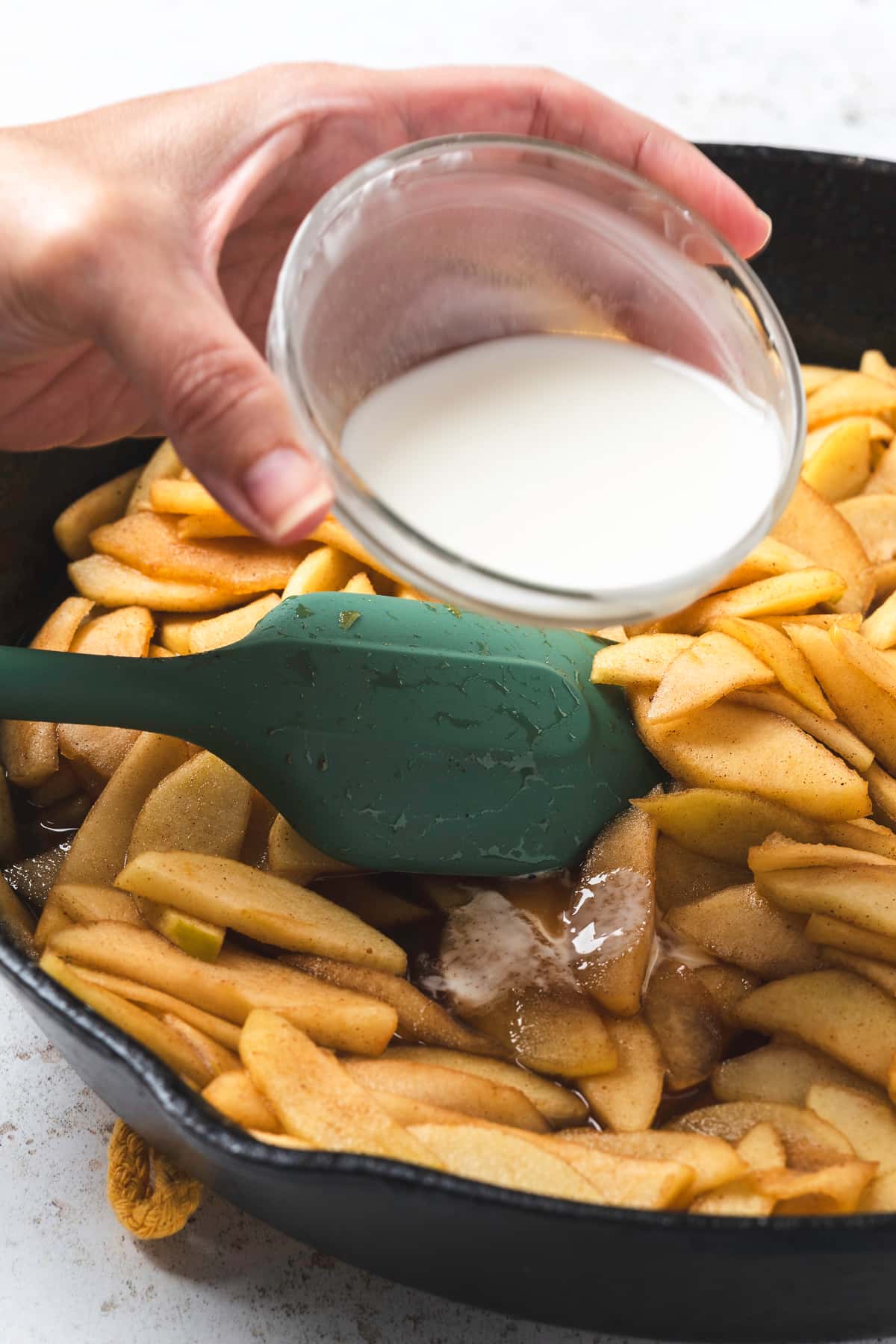 Hand pouring cornstarch mixture into a skillet of cooking apples with a spatula.