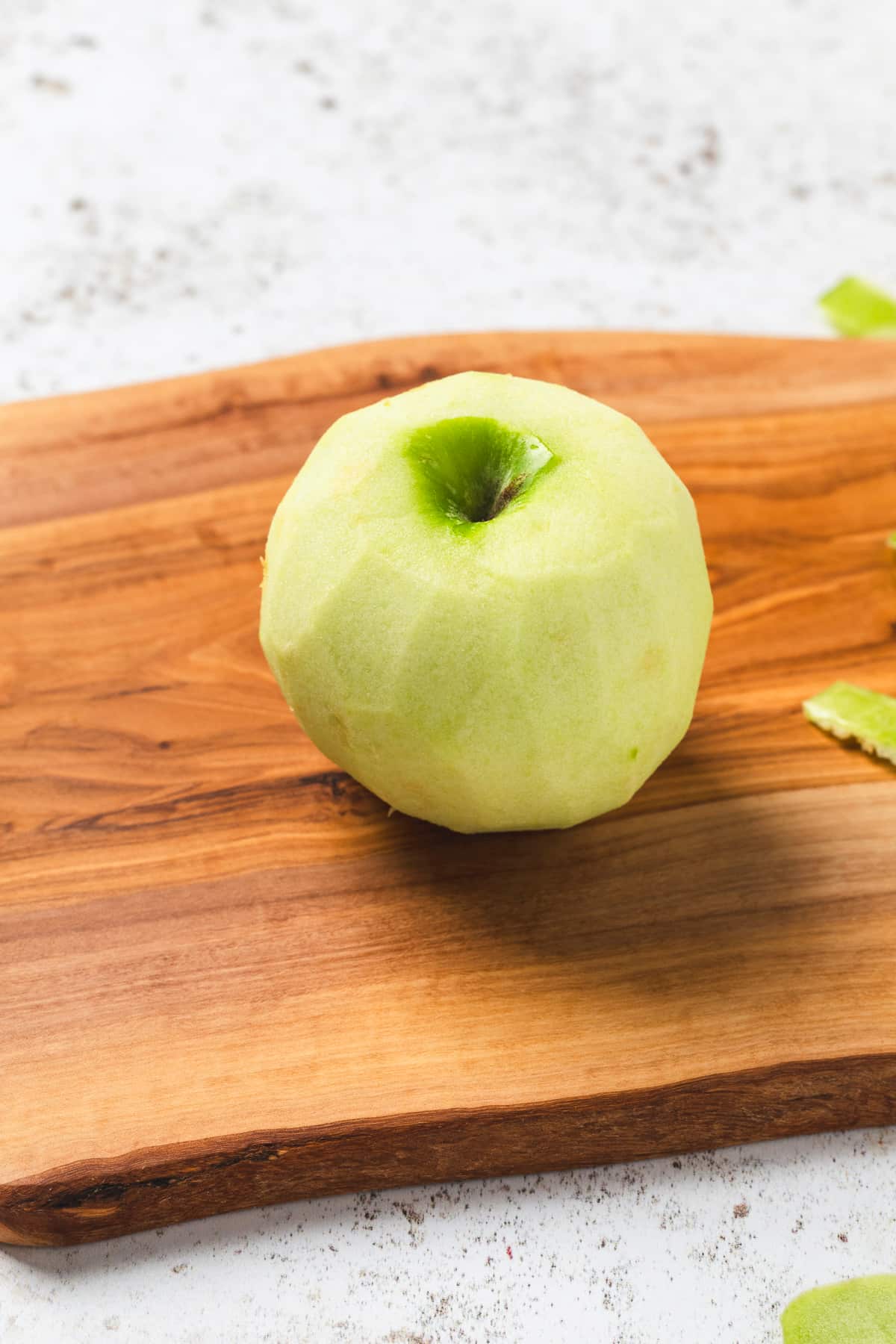 Peeled green apple on a wooden cutting board with bits of apple peel nearby.