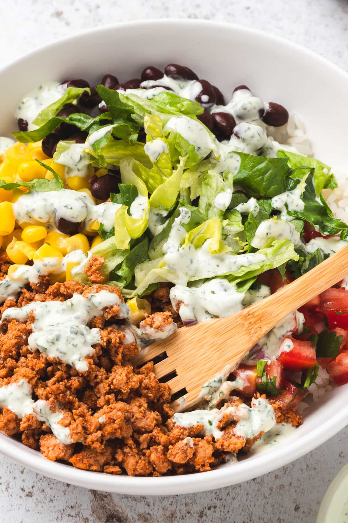 Turkey taco bowl with rice, beans, corn, lettuce, pico de gallo, and creamy dressing, wooden fork in bowl.