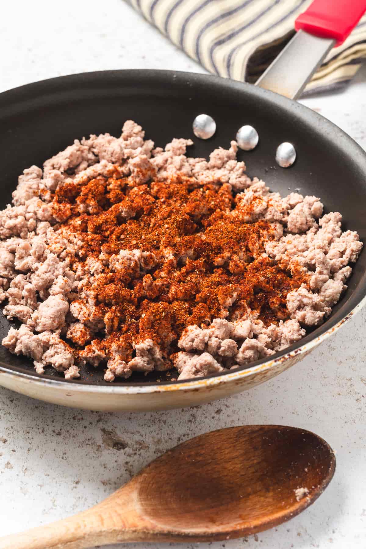 Ground turkey cooking in skillet with taco seasoning sprinkled on top, wooden spoon beside pan.