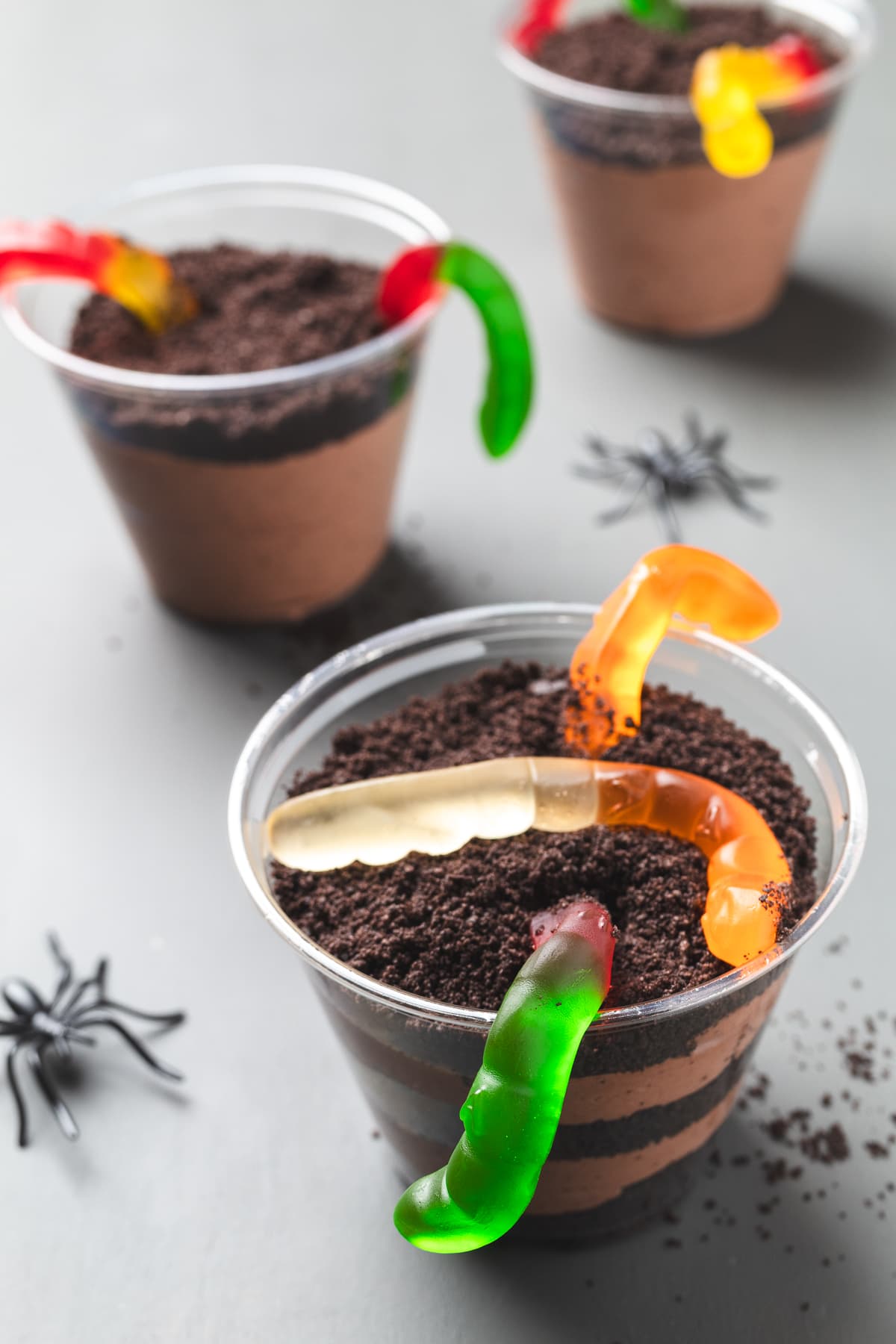 Multiple dirt cups layered with Oreo crumbs and pudding, decorated with gummy worms and toy spiders.