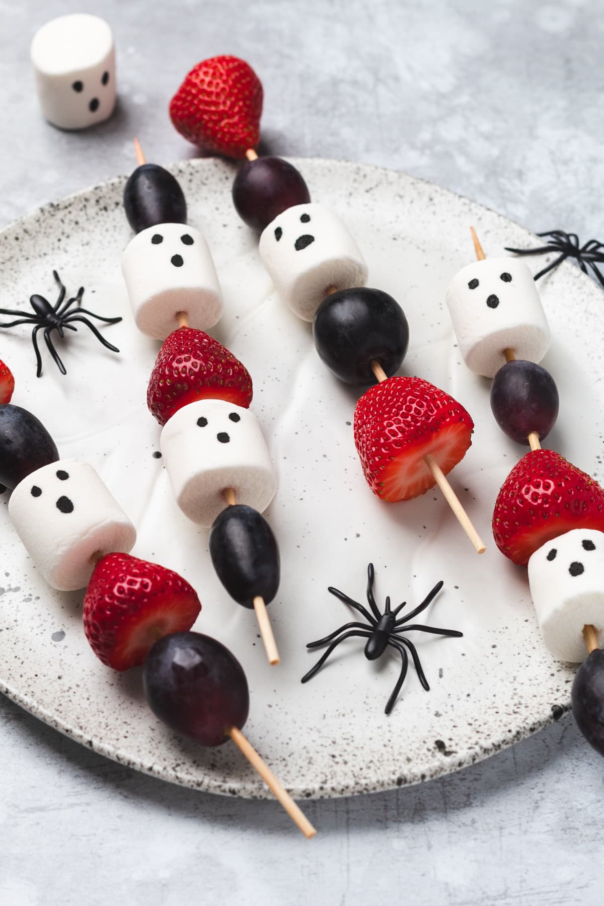 Skewers with marshmallow ghosts, strawberries, and grapes arranged on a plate with plastic spiders.