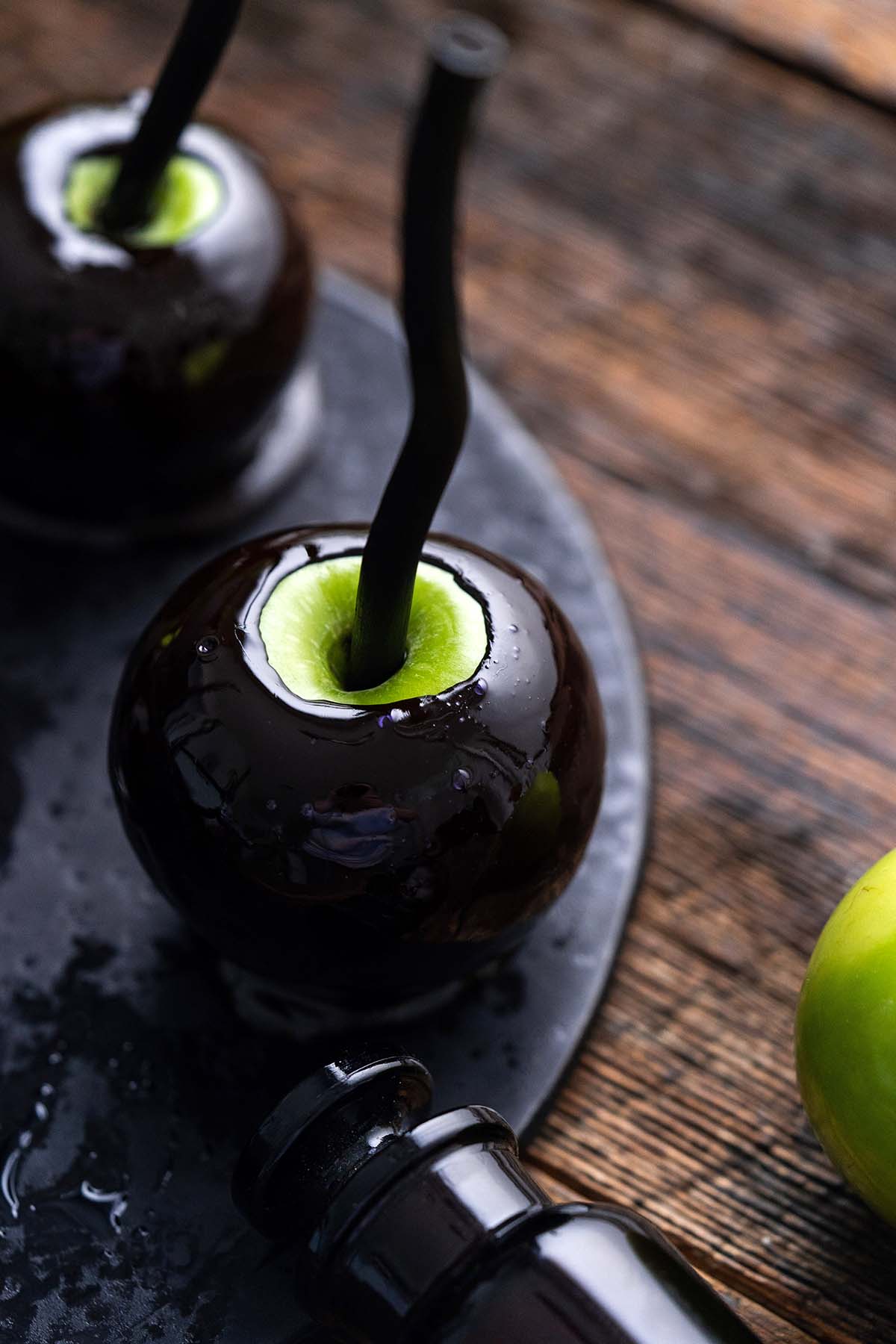 Glossy black candy apples with green centers and black straws on a rustic wooden table.