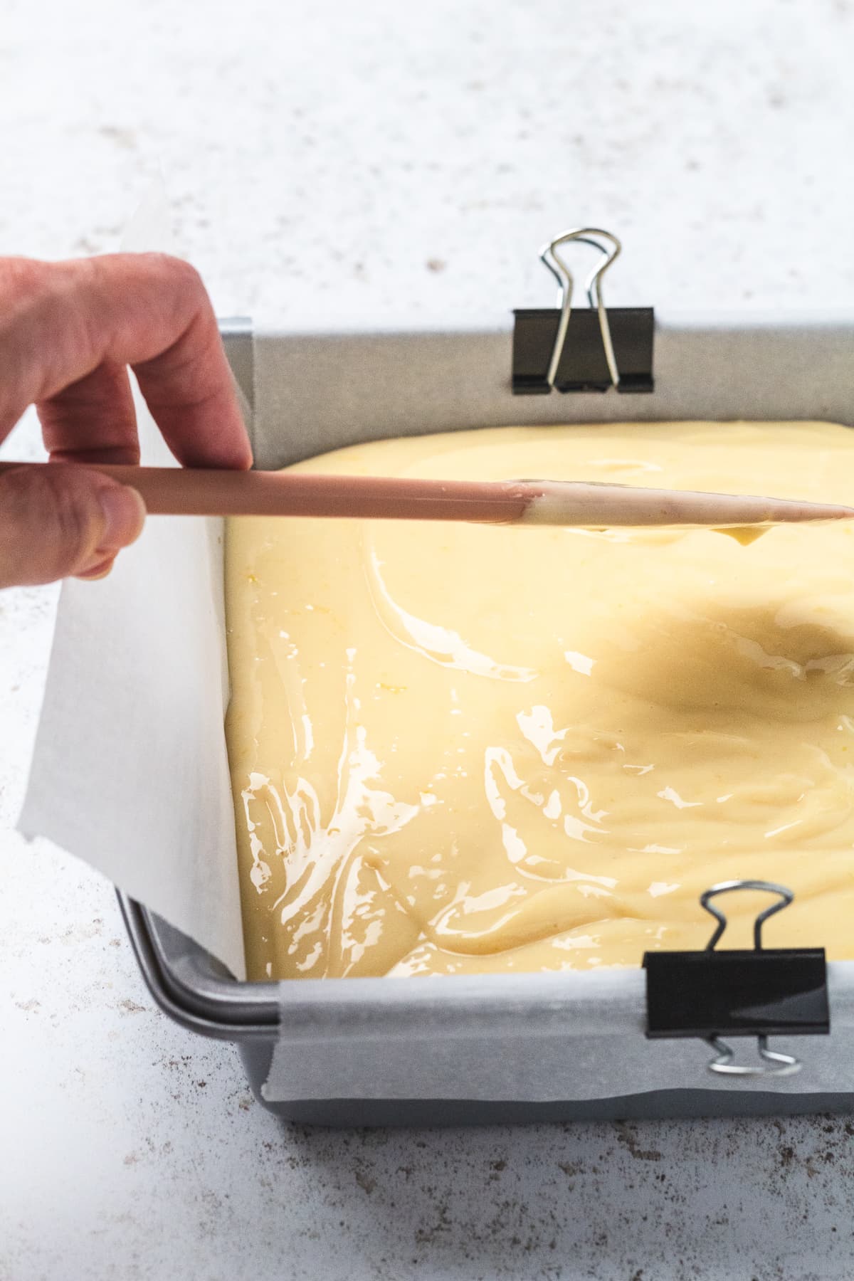 Spreading creamy lemon filling into a parchment-lined pan with a spatula.