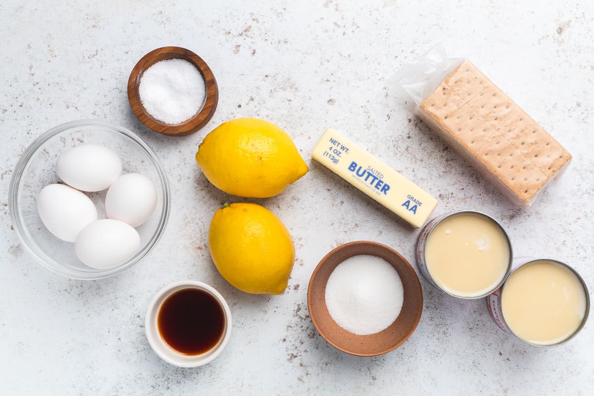 Lemons, eggs, butter, sugar, condensed milk, graham crackers, and vanilla arranged on counter.
