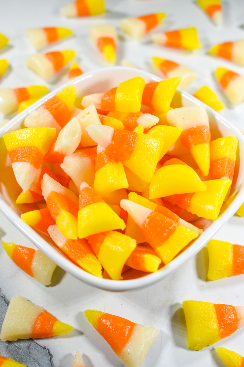 Bowl of homemade candy corn pieces in yellow, orange, and white, surrounded by more candy corn on table.
