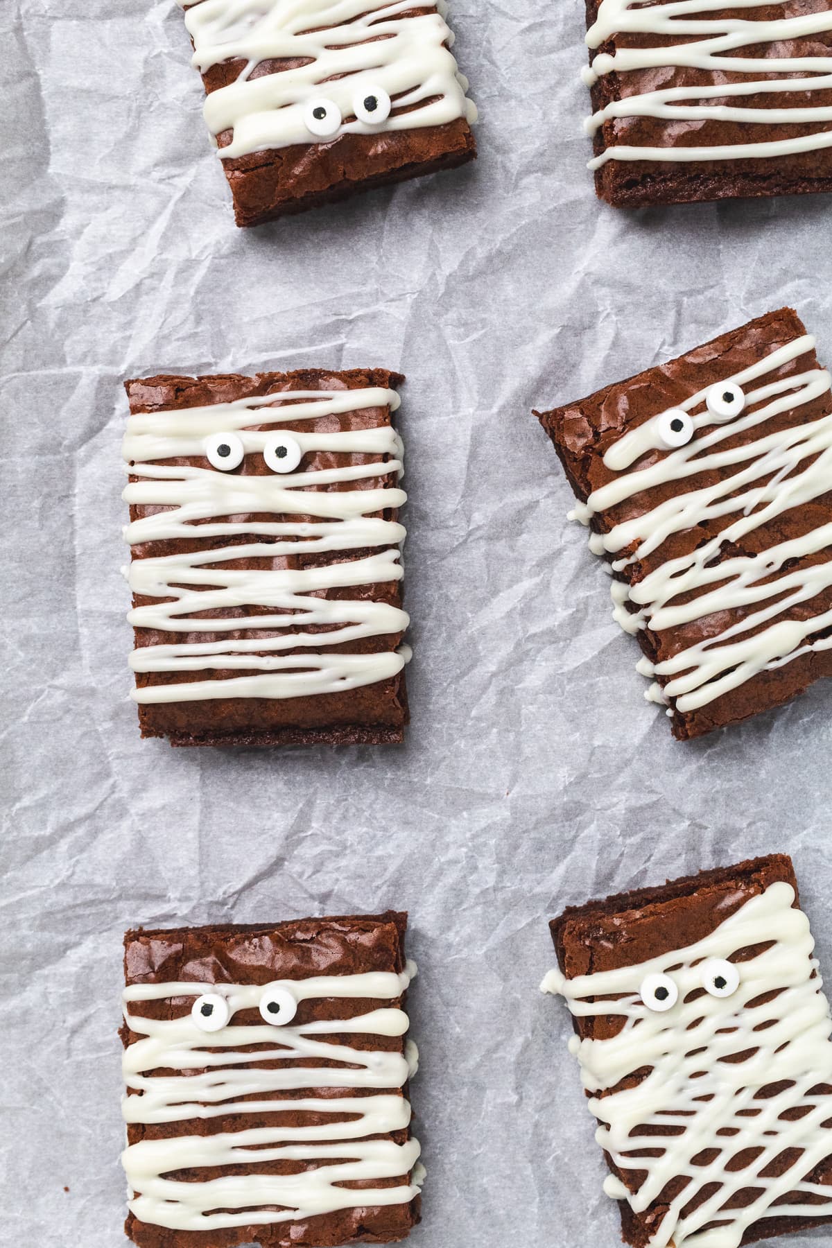 Mummy brownies topped with candy eyes and white chocolate drizzle on parchment paper.