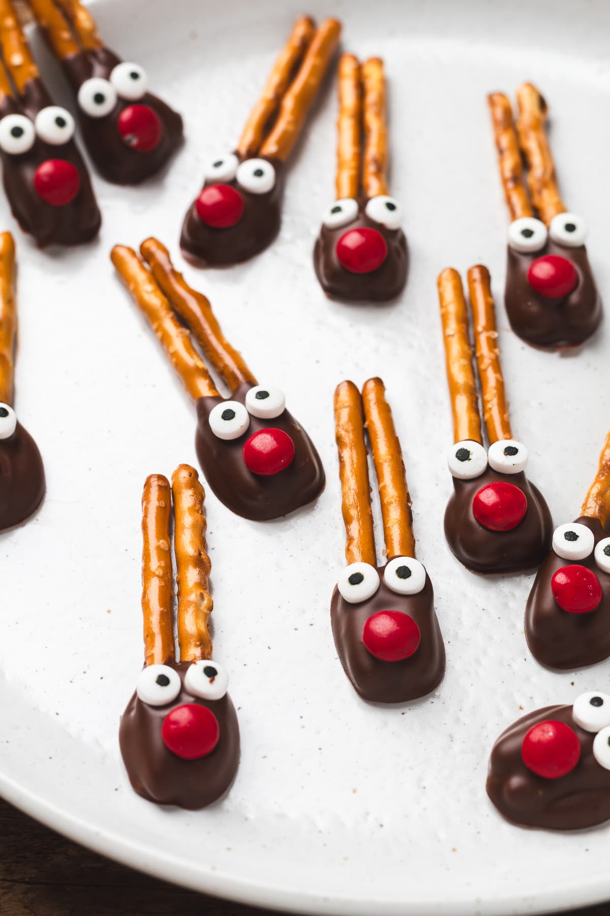 Chocolate-dipped pretzel reindeer with red candy noses and candy eyes on a white plate.