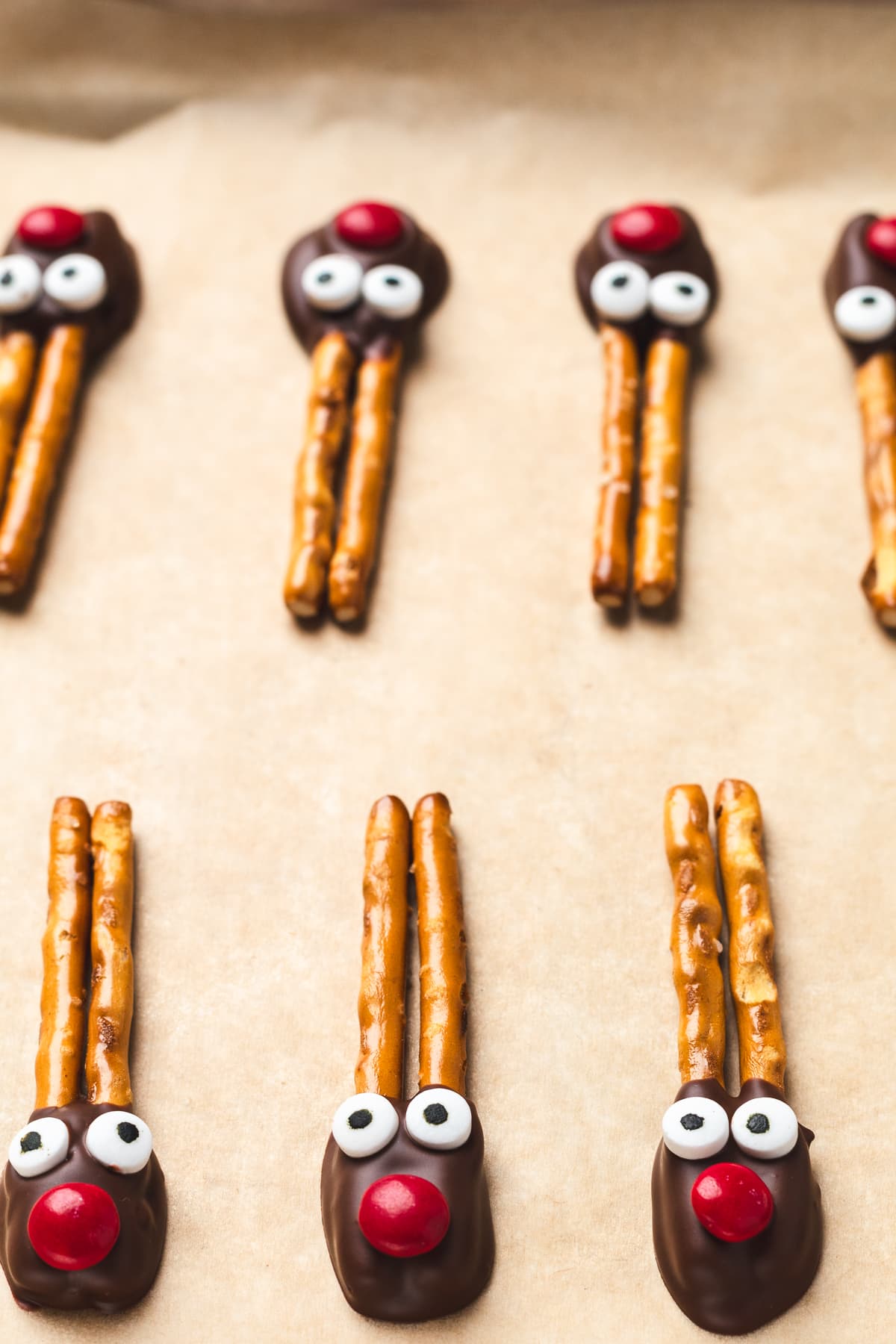 Reindeer pretzels with chocolate faces, candy eyes, and red noses arranged neatly on parchment paper.