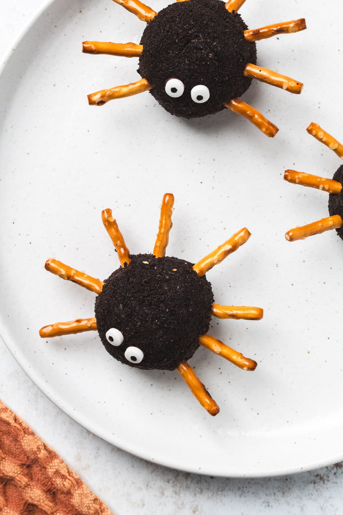 Oreo-coated spider cheeseballs with candy eyes and pretzel legs arranged on a white plate.