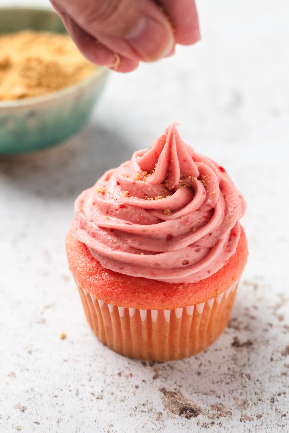 Hand sprinkling graham cracker crumbs over a strawberry cheesecake cupcake.