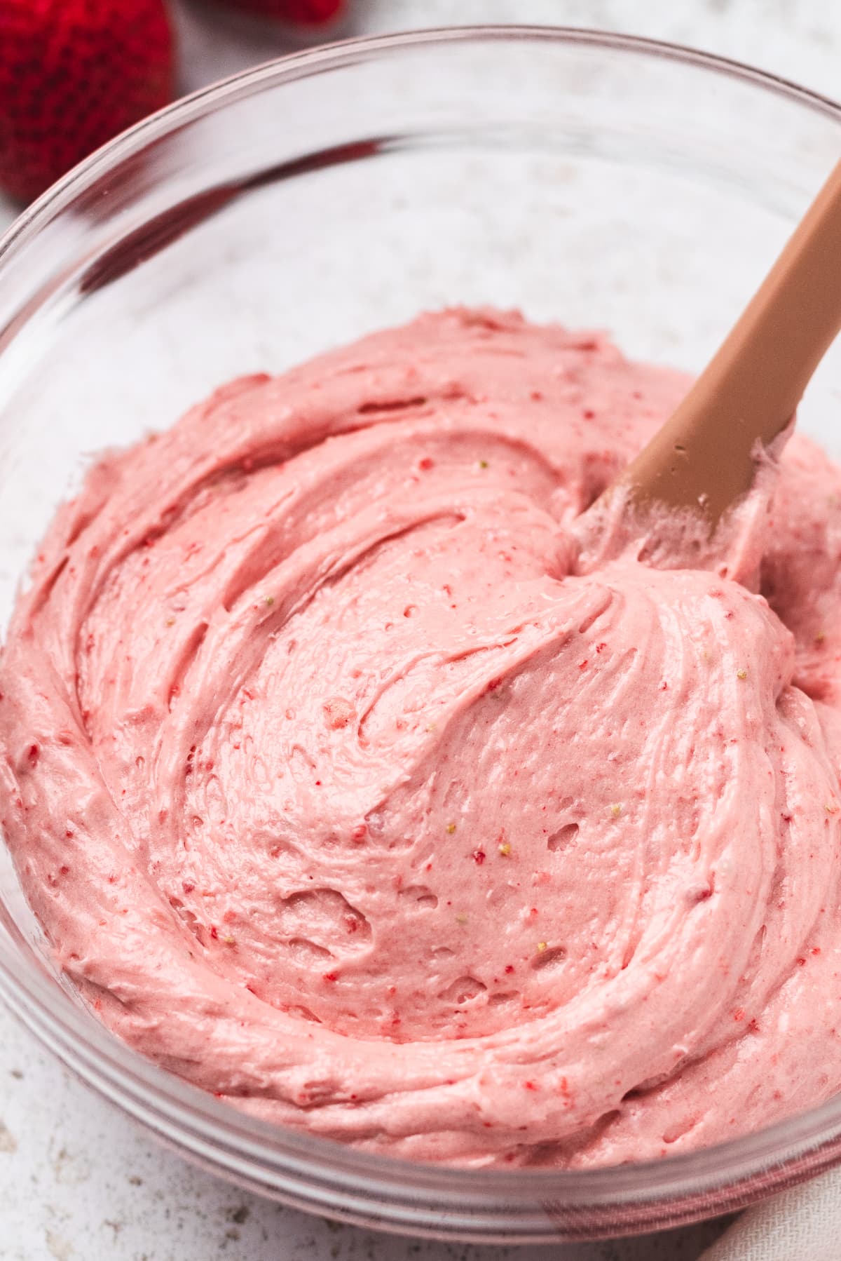 Strawberry cheesecake frosting in a glass bowl with a spatula, smooth and creamy with specks of strawberry.