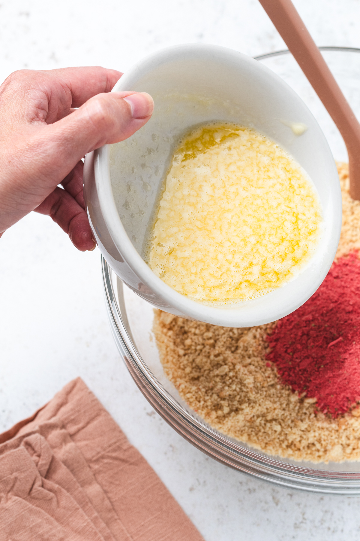 Hand holding a bowl pouring butter into a bowl filled with pulverized cookies and freeze-dried strawberries.