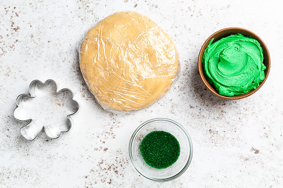 Cookie dough wrapped in plastic with green frosting, sprinkles, and Shamrock cutter nearby.