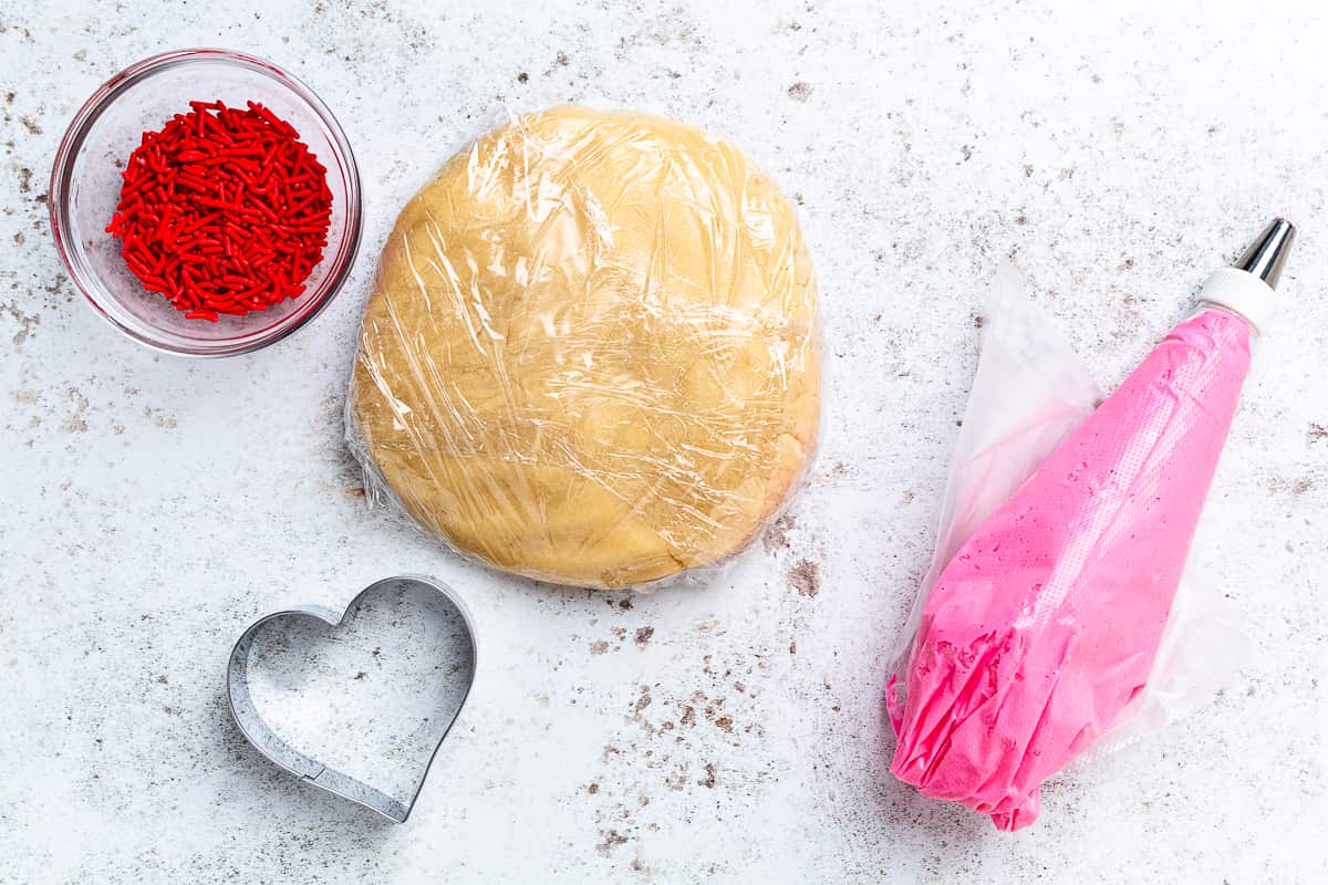 Sugar cookie dough with heart cookie cutter, red sprinkles, and piping bag of pink frosting.