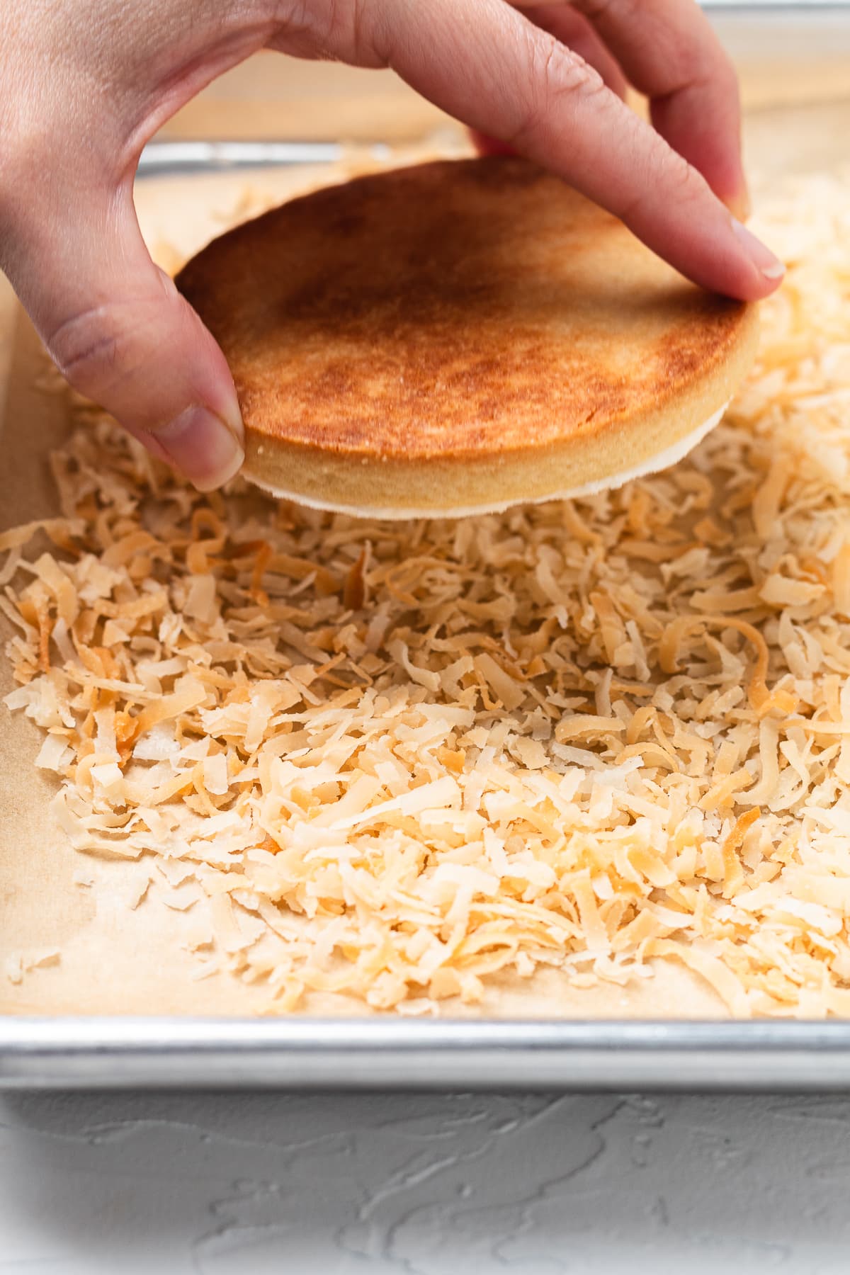 Hand pressing a frosted cookie onto toasted coconut on a baking sheet to coat the top.