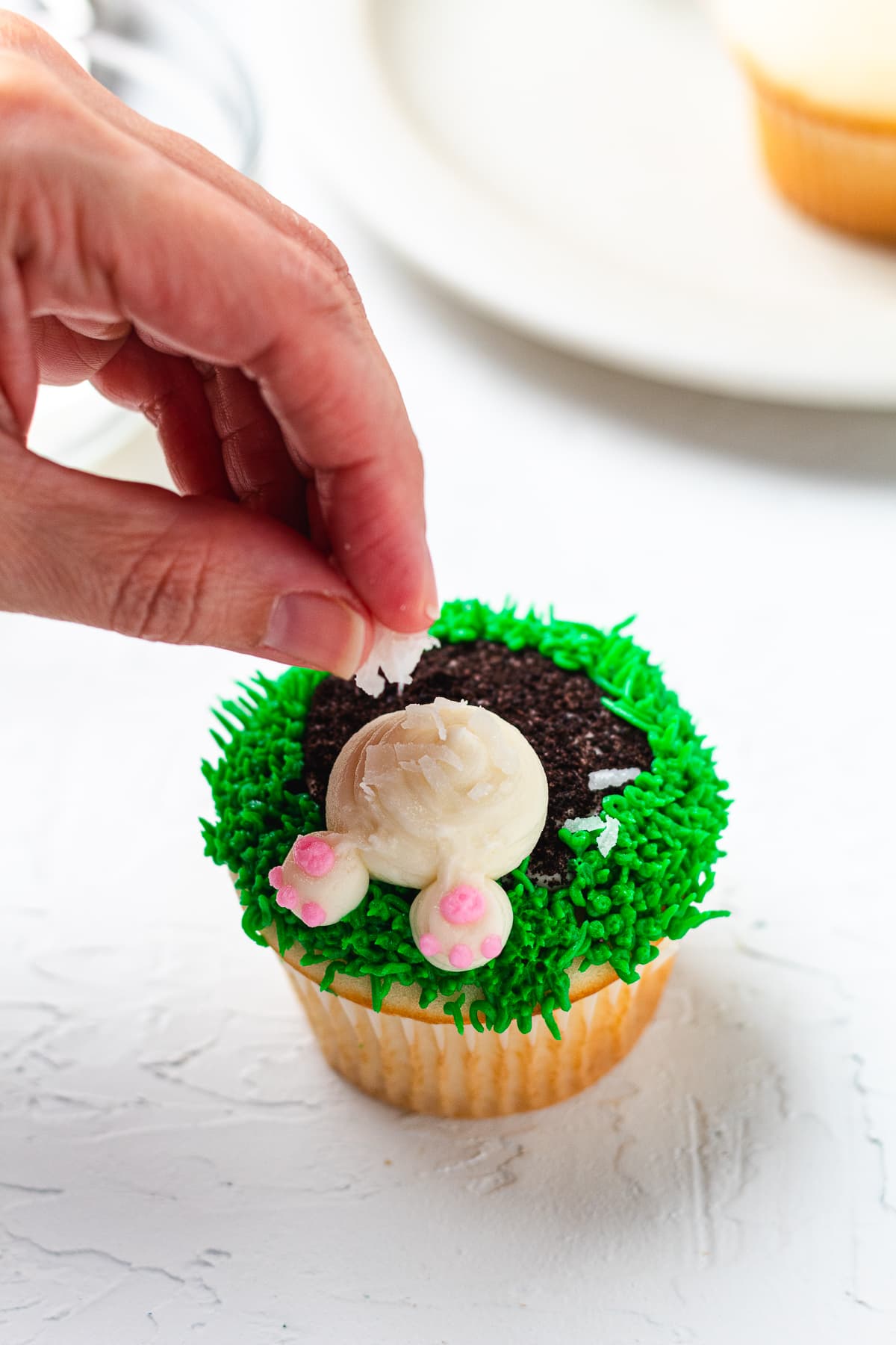 Hand sprinkling shredded coconut onto the bunny tail frosting on a decorated cupcake.