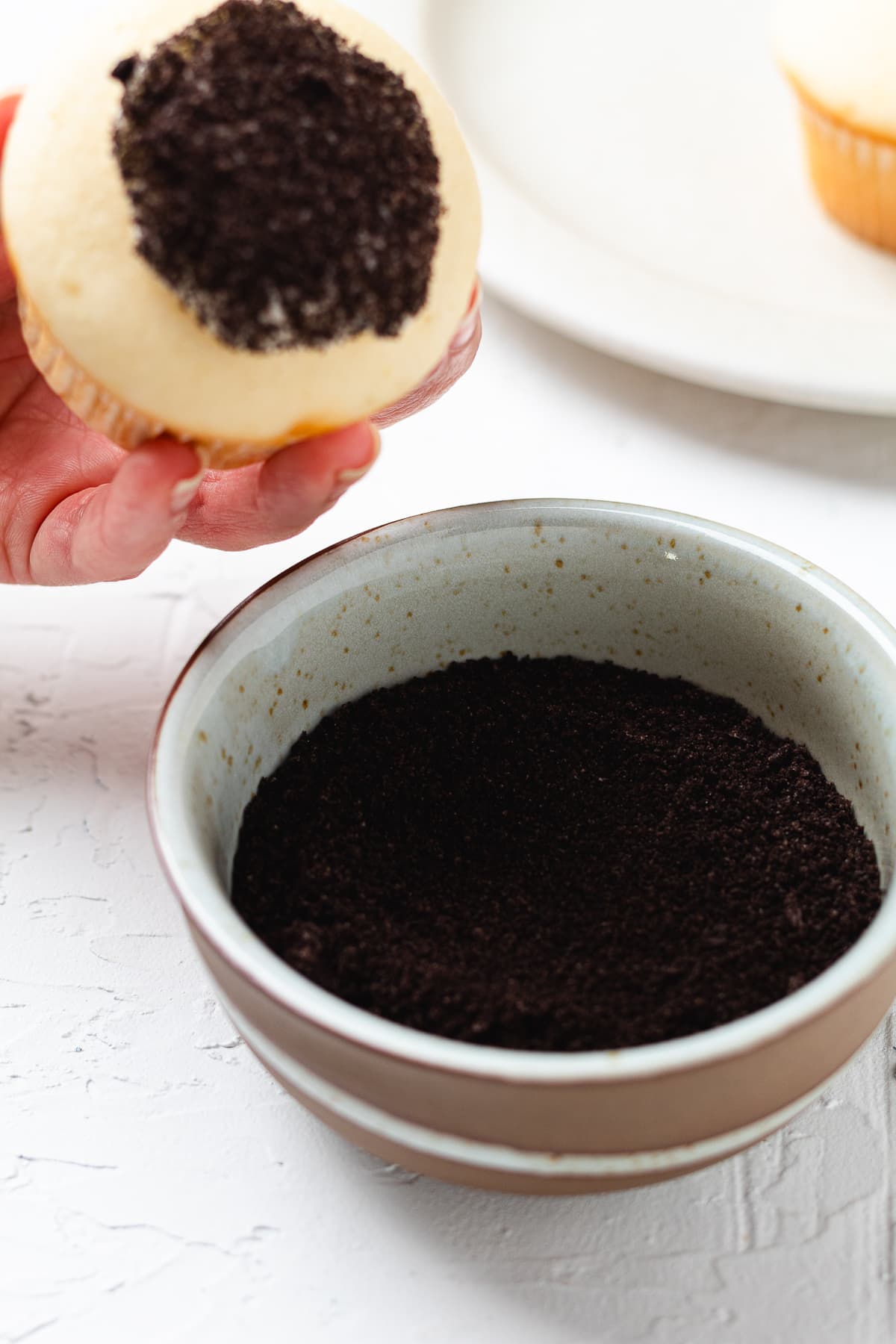 Hand pressing the top of a frosted cupcake into chocolate cookie crumbs to coat it.