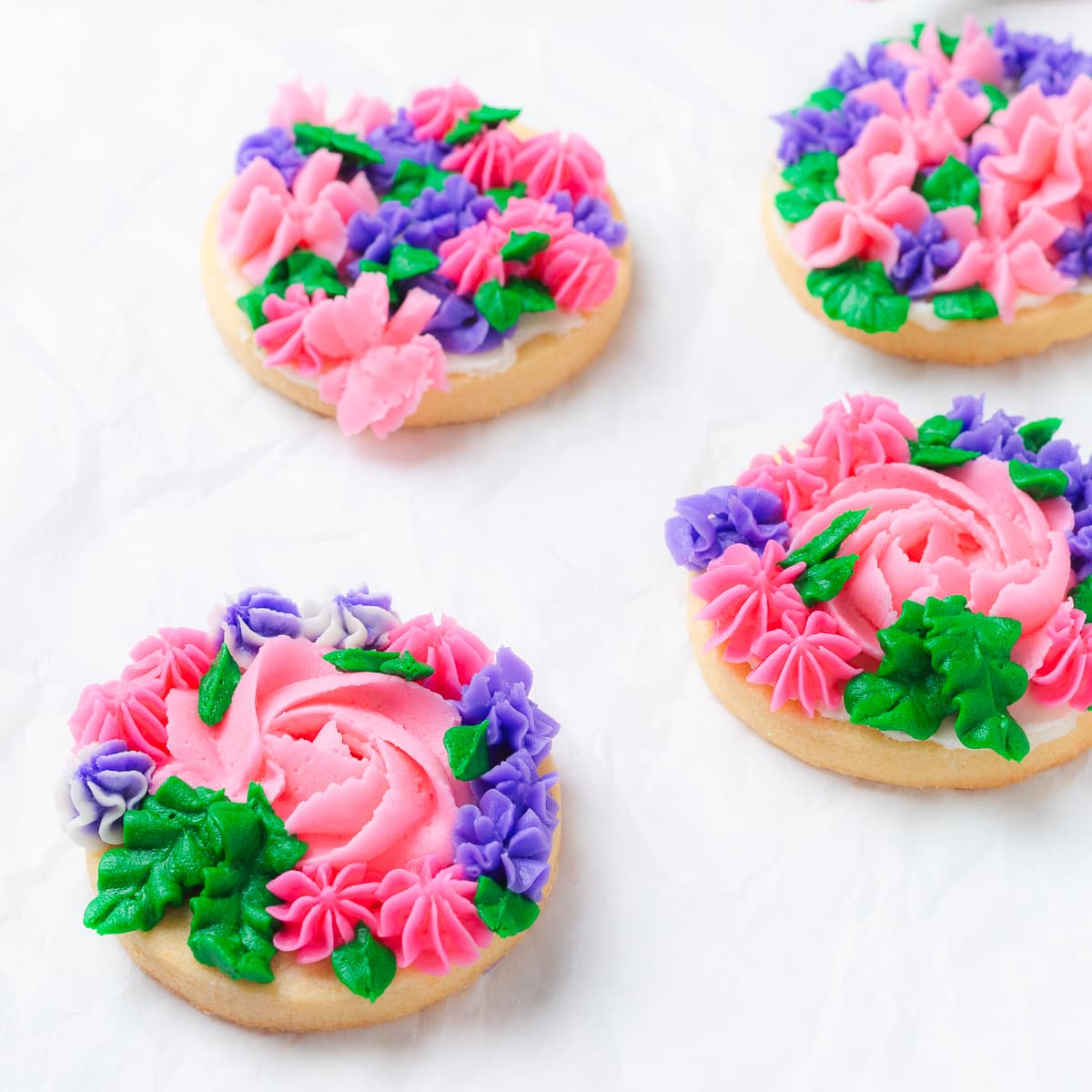 Close-up of decorated flower cookies with pink and purple buttercream roses and green leaves.