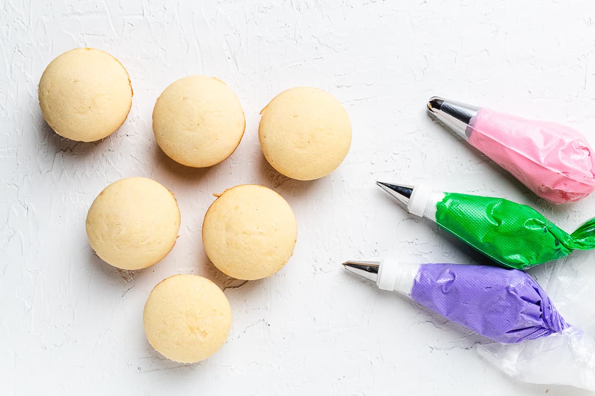 Plain cupcakes with piping bags filled with pink, purple, and green frosting.