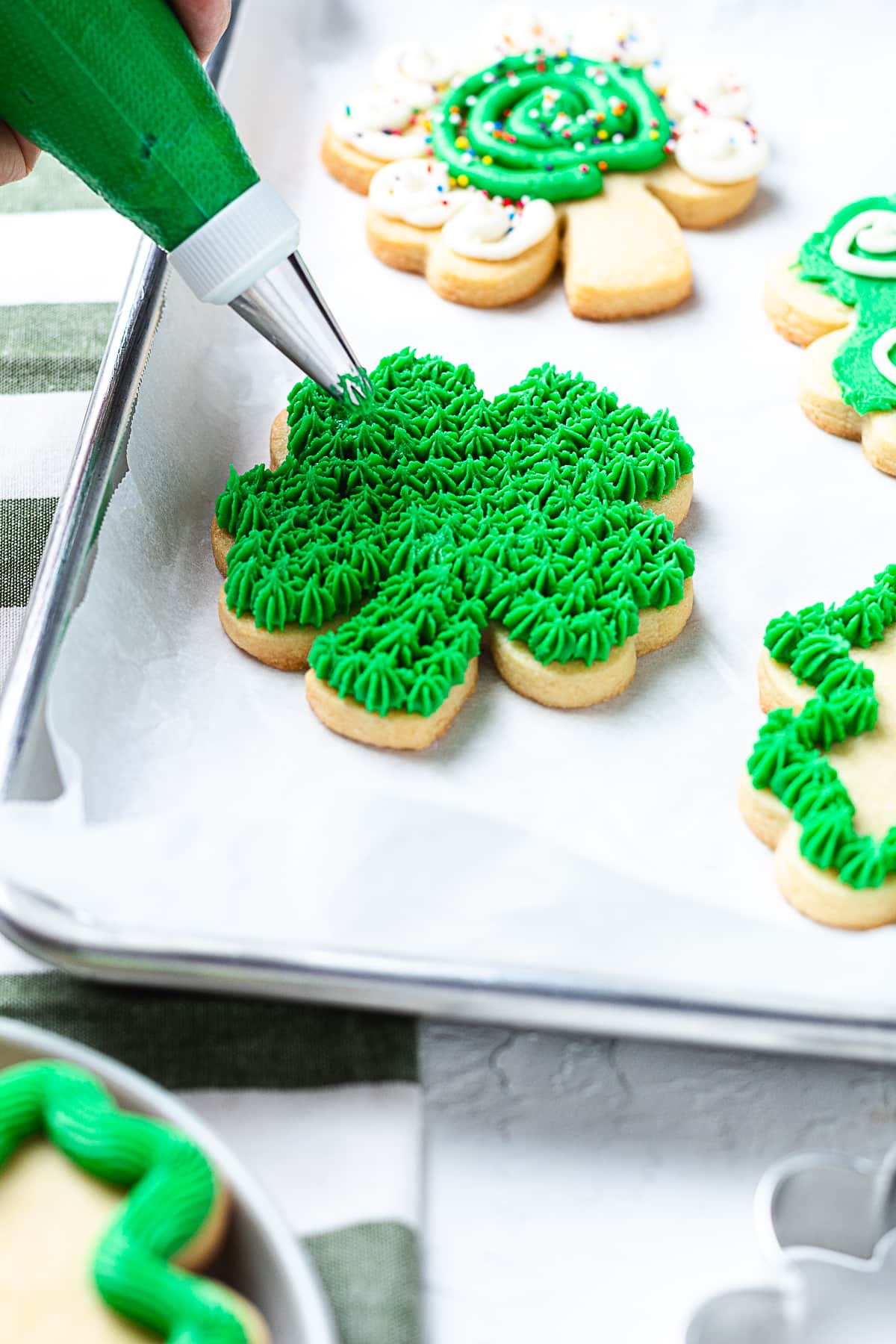 Piping green frosting onto shamrock sugar cookie with star tip on parchment-lined tray.