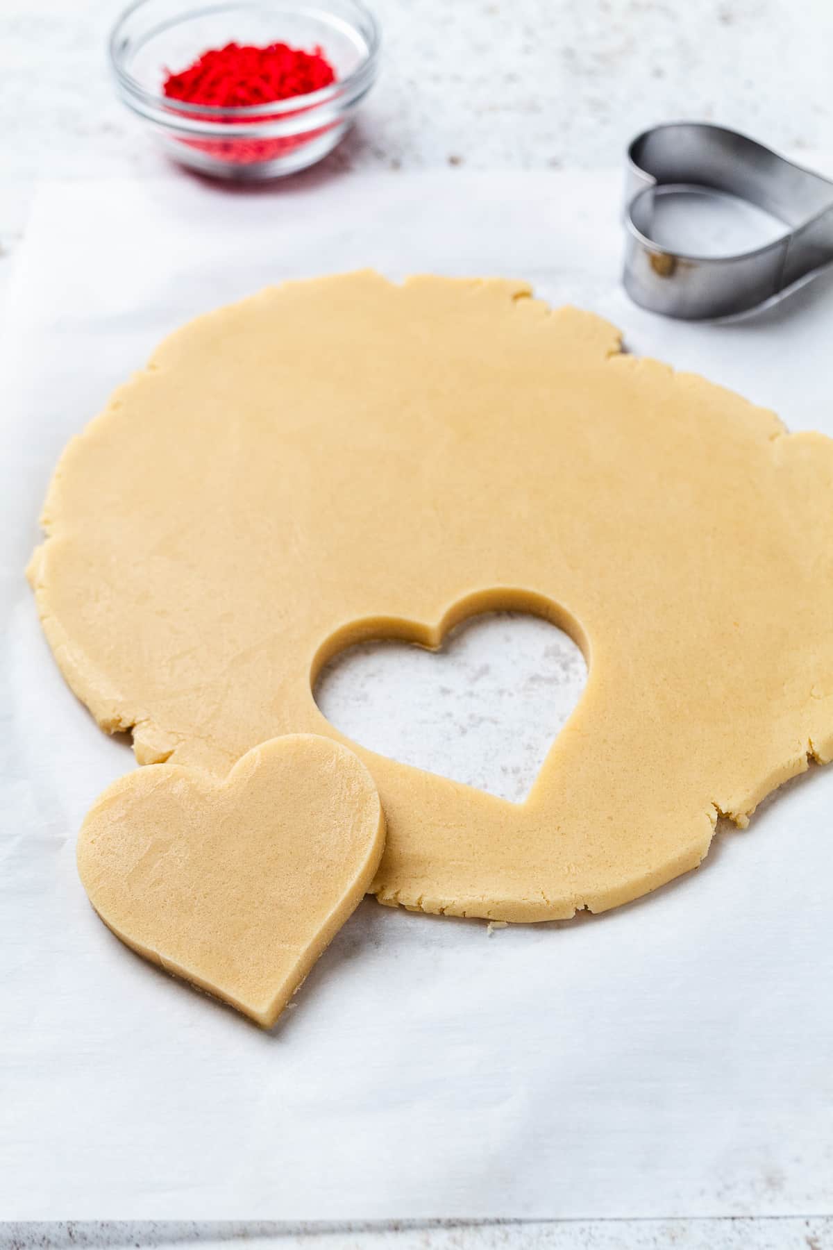 Heart cookie shape being cut from rolled sugar cookie dough with cutter nearby.
