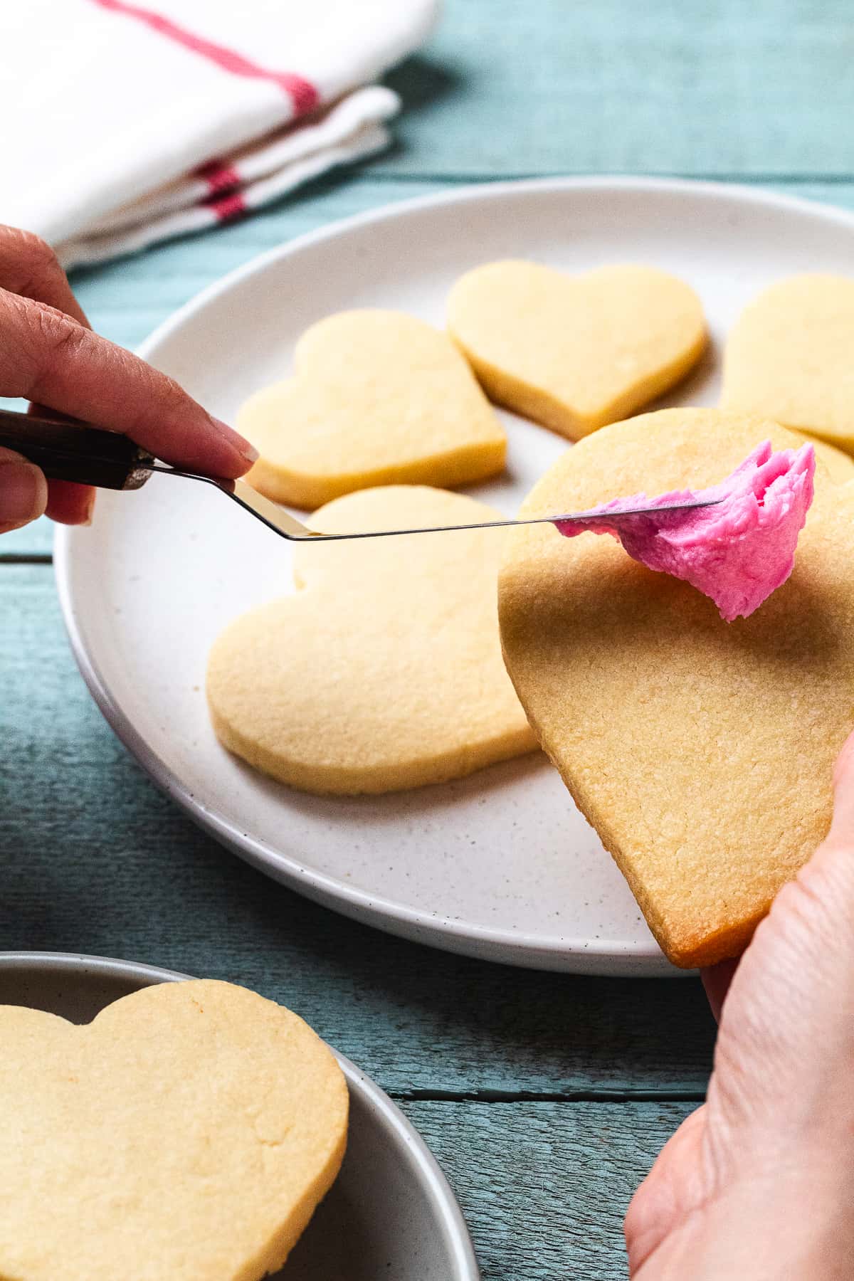 Spreading pink frosting onto heart-shaped sugar cookies on a plate.