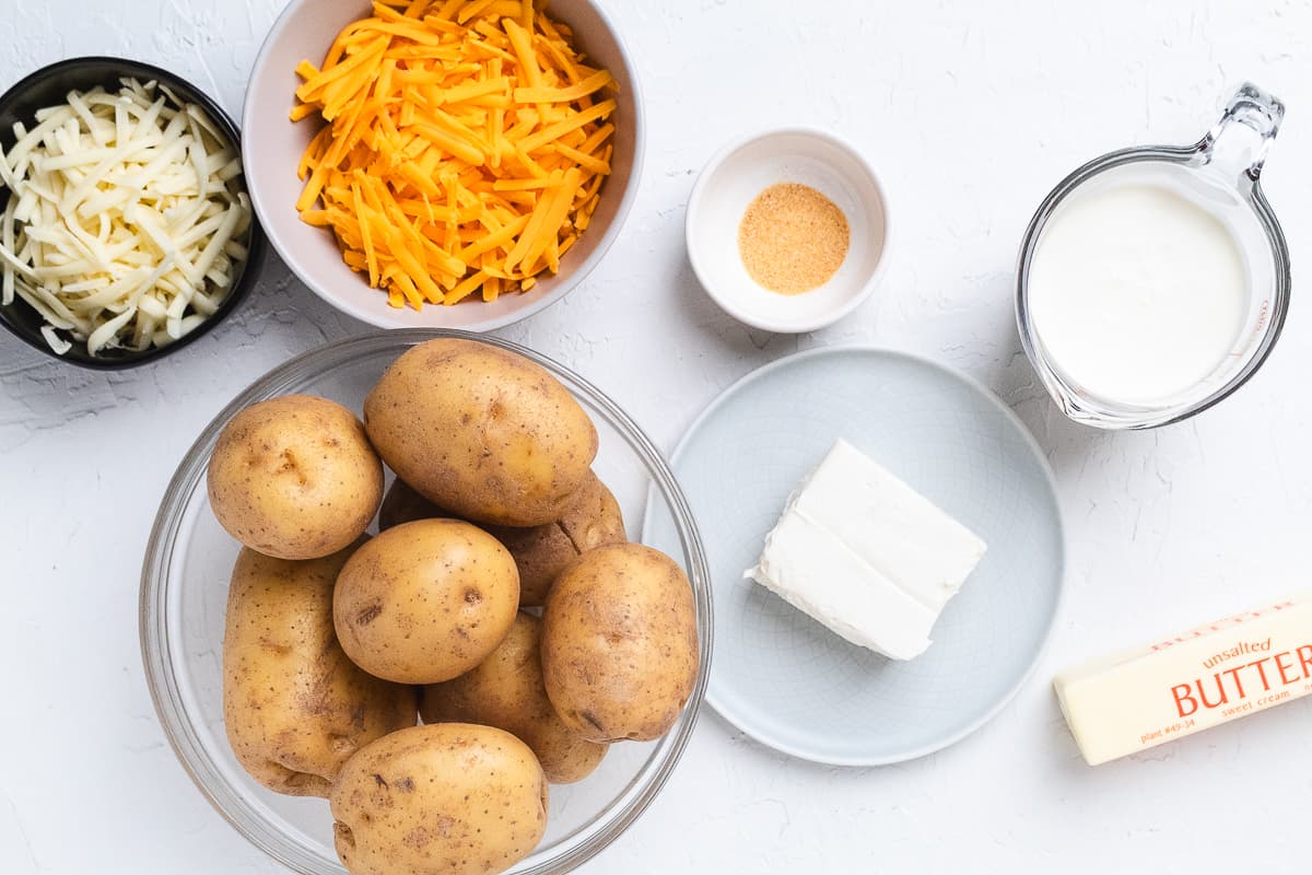 Ingredients for cheesy mashed potatoes including Yukon gold potatoes, shredded cheddar, shredded mozzarella, cream cheese, butter, heavy cream, and seasoning displayed on a white surface.