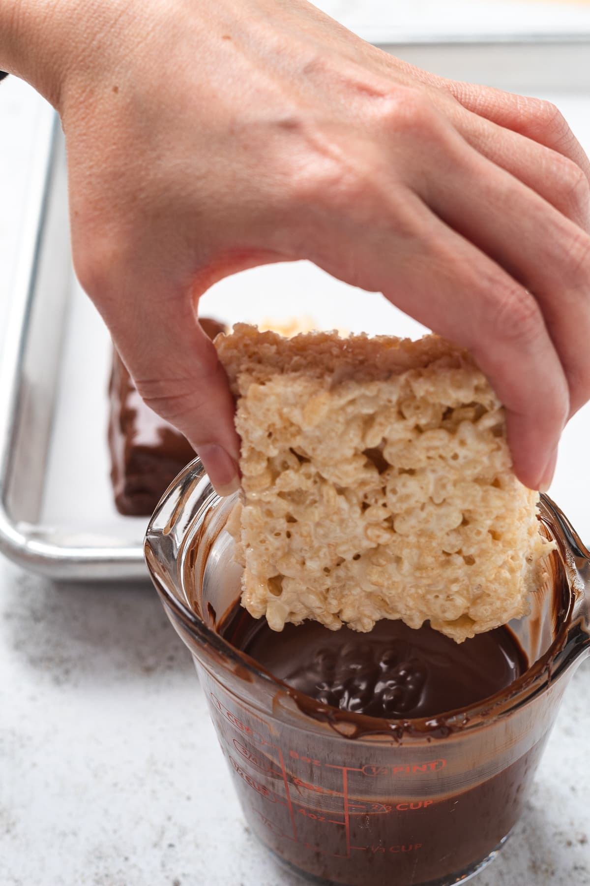 Hand dipping a Rice Krispie square into melted chocolate in a glass measuring cup.