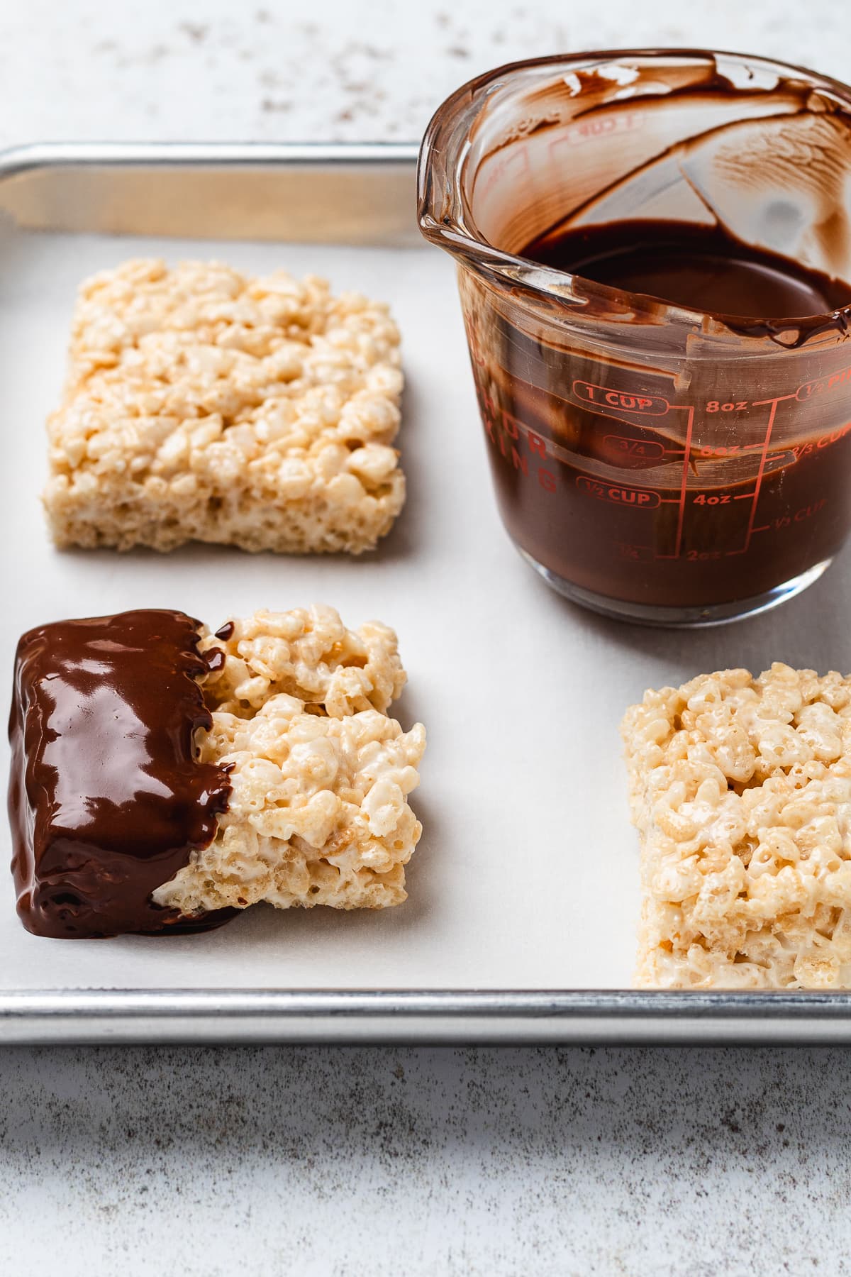 Freshly dipped Rice Krispie treats setting on a parchment-lined baking sheet beside melted chocolate.