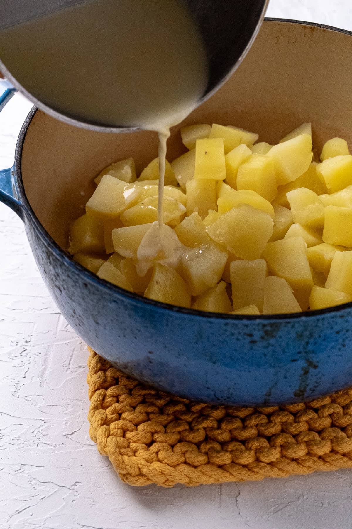 Warm cream mixture being poured over cooked potato chunks in a blue pot.