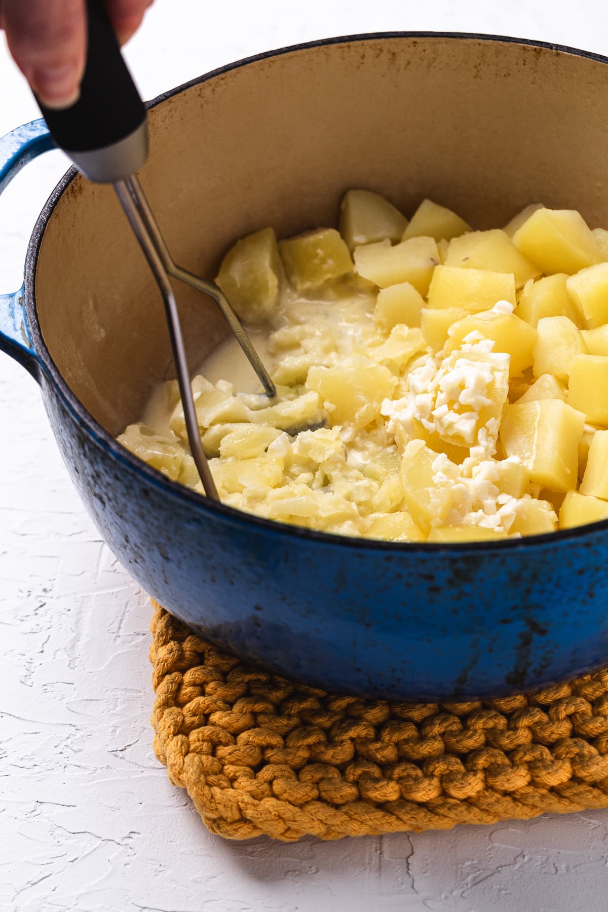 Hand mashing cooked potatoes with cream and butter in a large blue pot.
