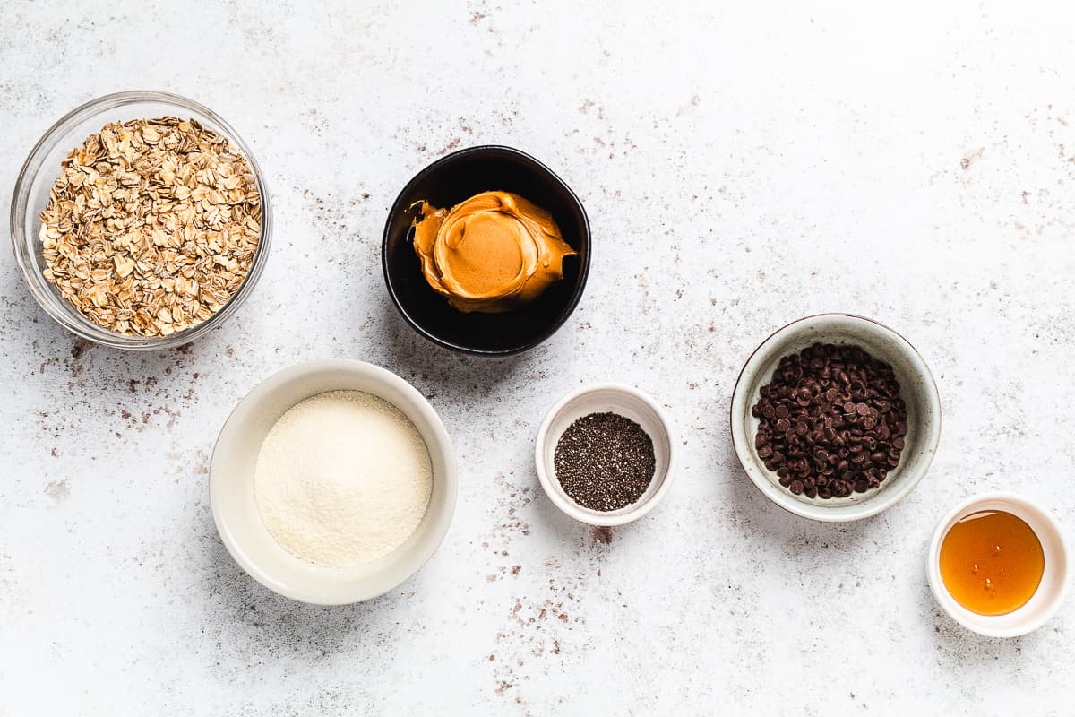 Overhead view of ingredients for protein balls laid out in small bowls on a white surface.