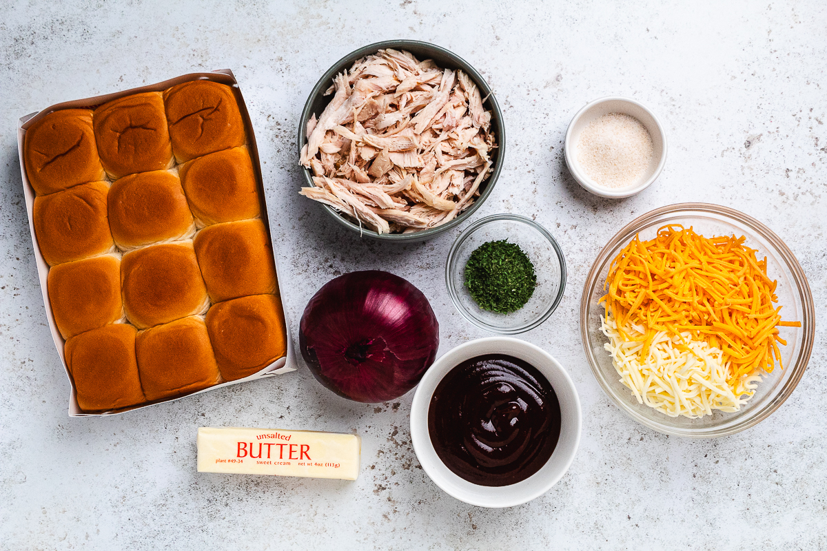 Ingredients for BBQ chicken sliders arranged on a table, including rolls, chicken, BBQ sauce, and cheese.