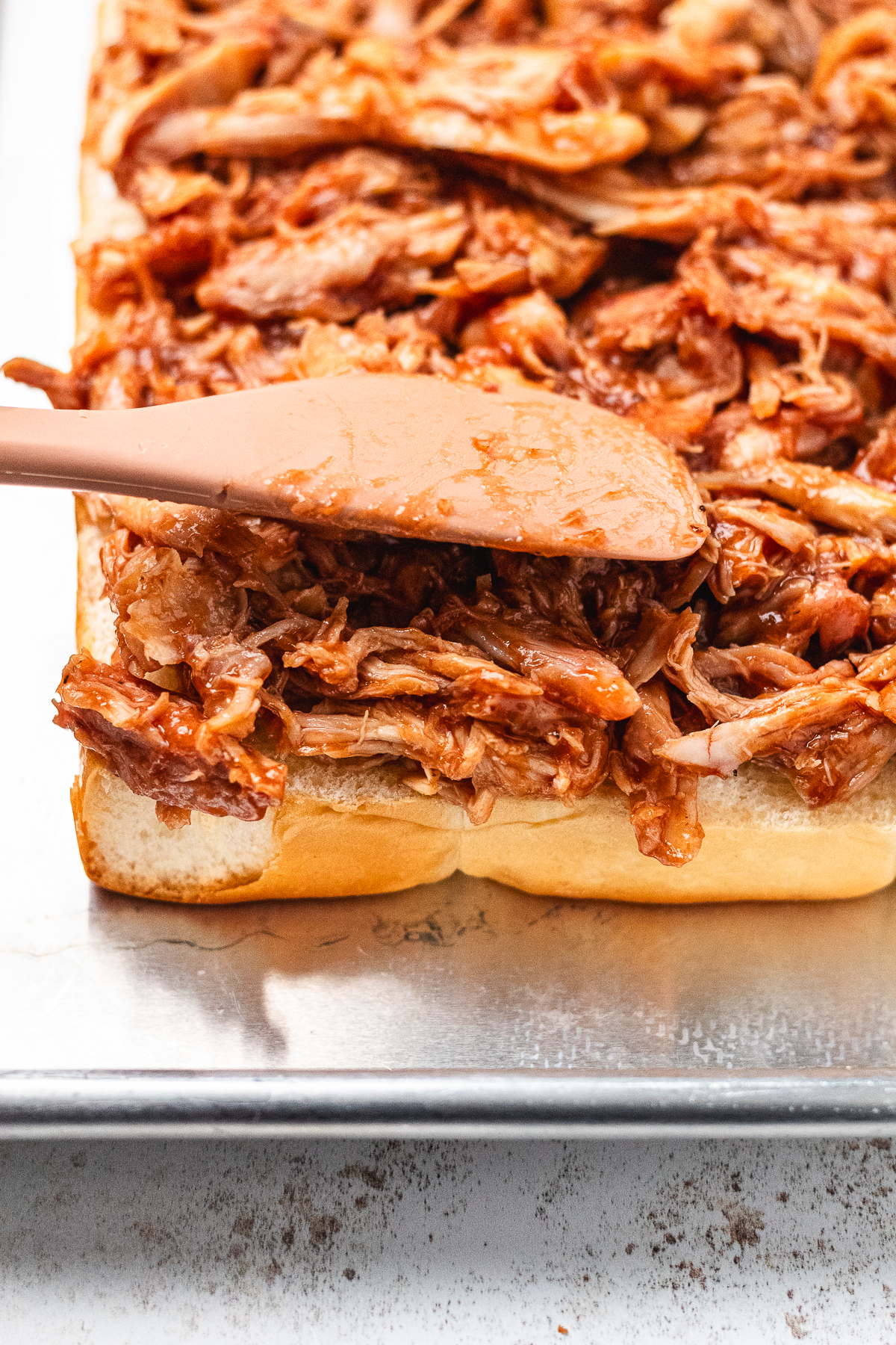 BBQ shredded chicken being spread evenly over sliced Hawaiian rolls before adding toppings.
